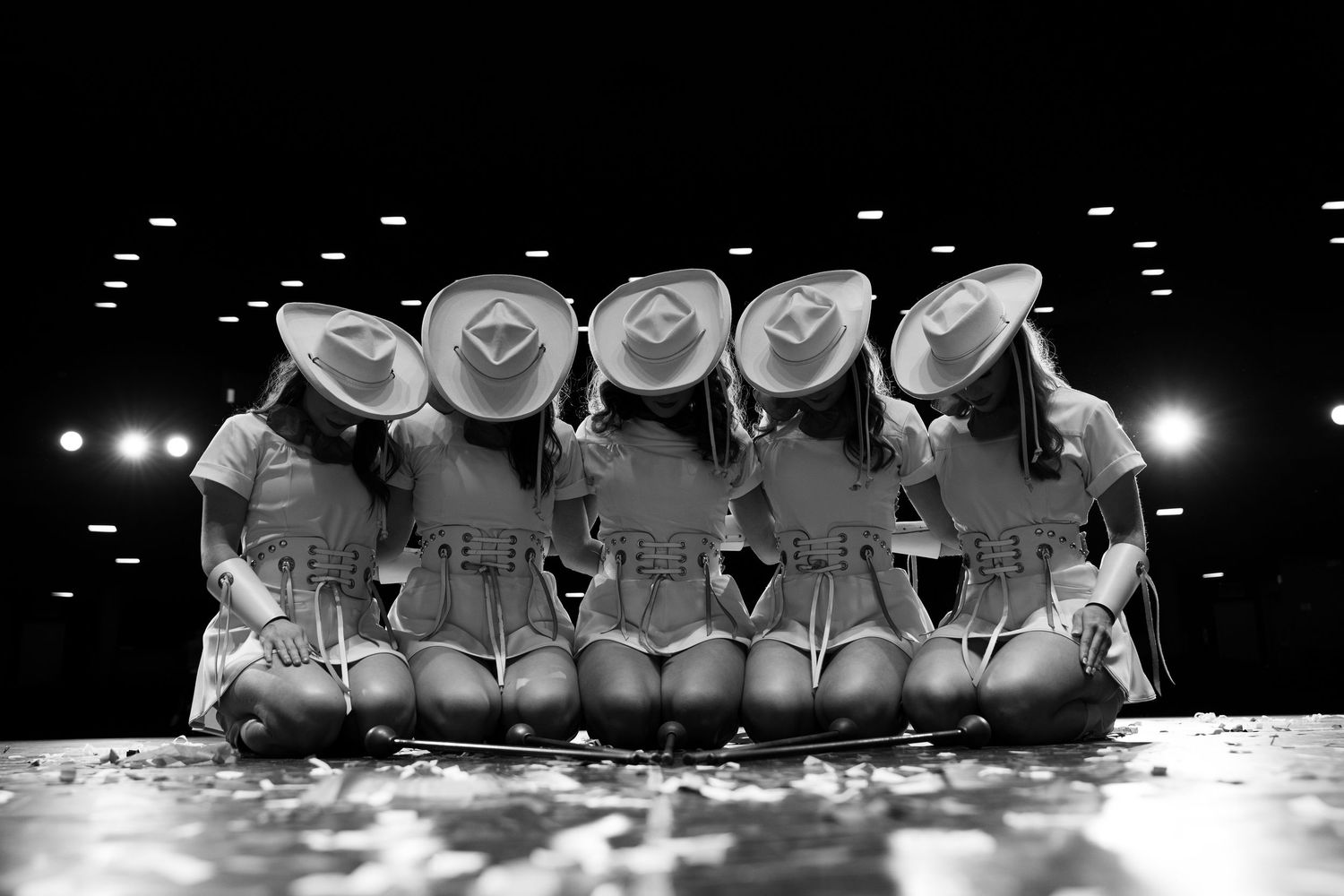Five girls in cowboy hats sit together on stage, viewed from behind, in a black and white performance photo.