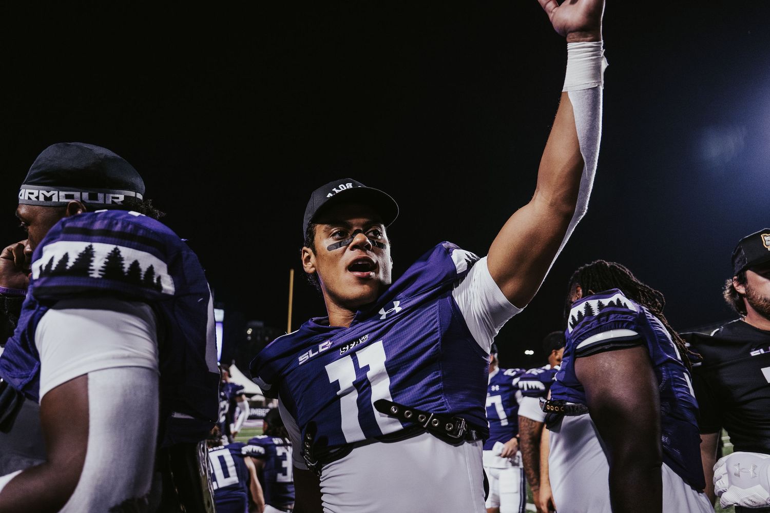 Football players celebrate on the field after a game with raised arms and excited gestures under stadium lights.