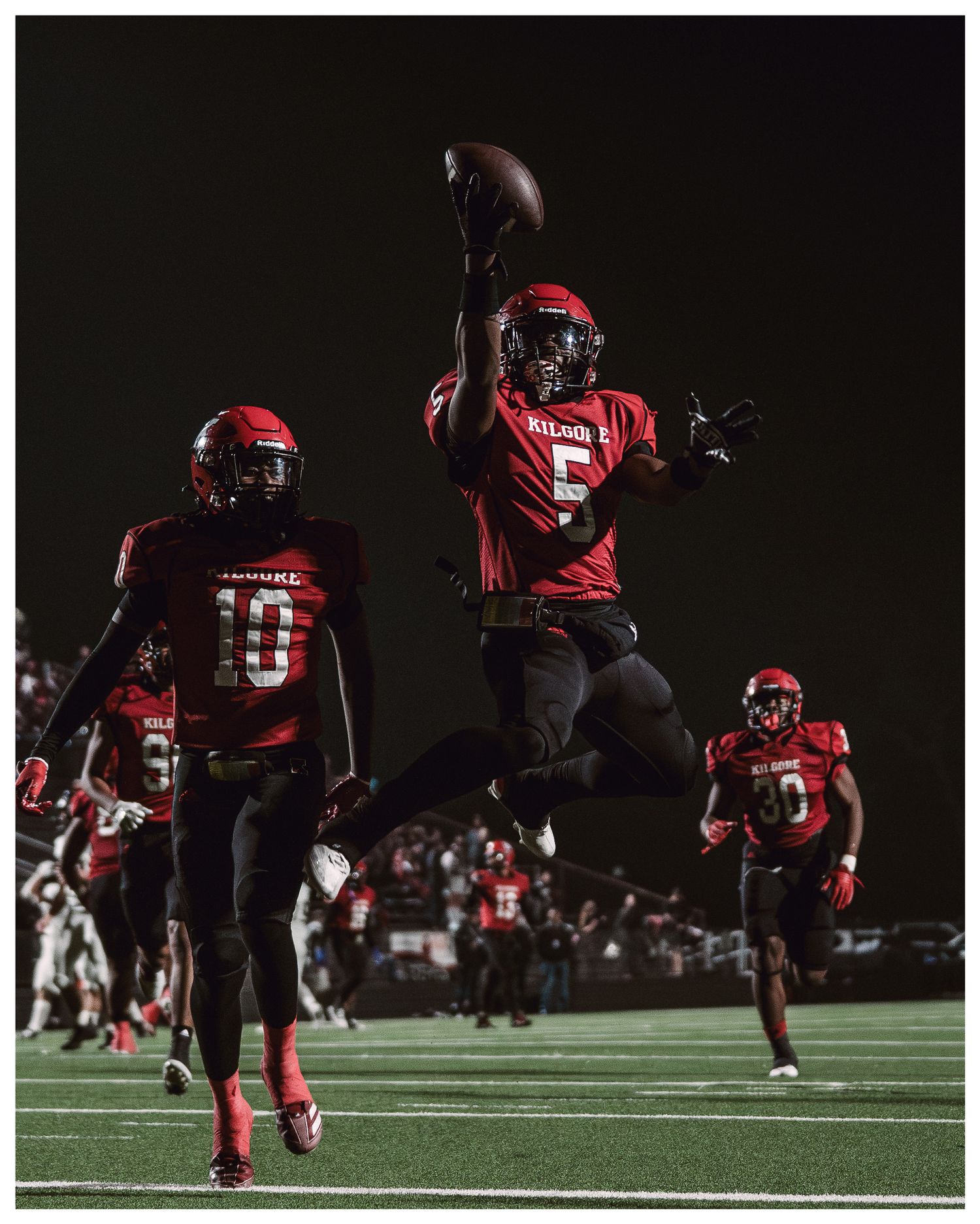 Football players in red uniforms perform dynamic jumping poses during nighttime practice.