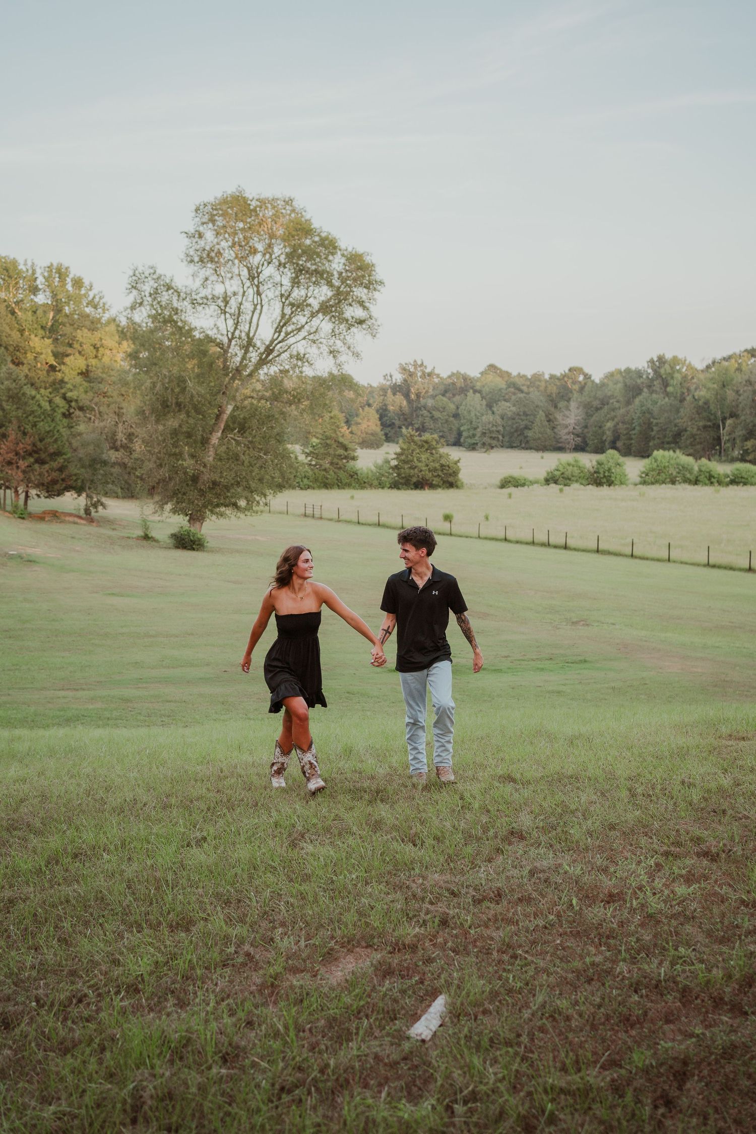 Couple holds hands while walking through scenic grassy field at sunset.