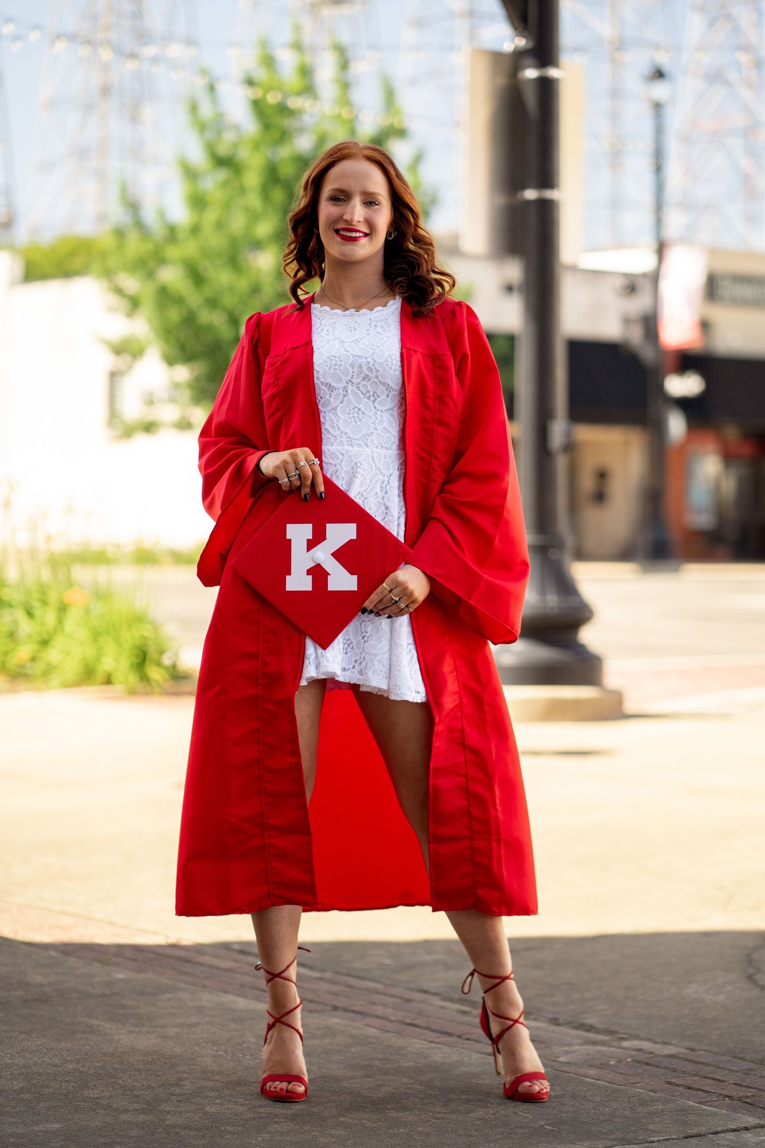 Person in red blazer and white lace top shows off letter K while posing on city sidewalk.