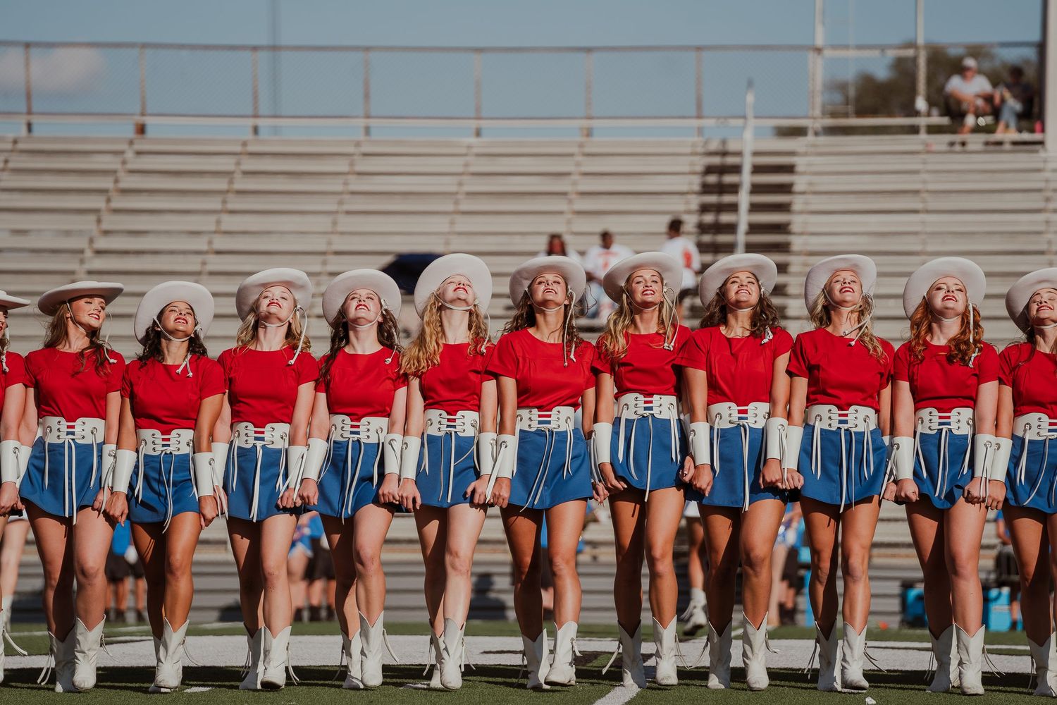 Cheerleaders in red and blue uniforms performing in a line formation at a stadium with spectators in the background.