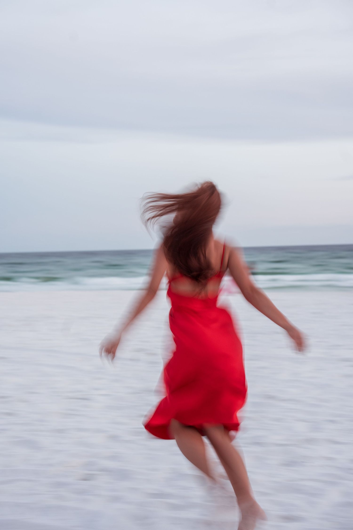 Woman in flowing red dress runs carefree along ocean shoreline at dusk.