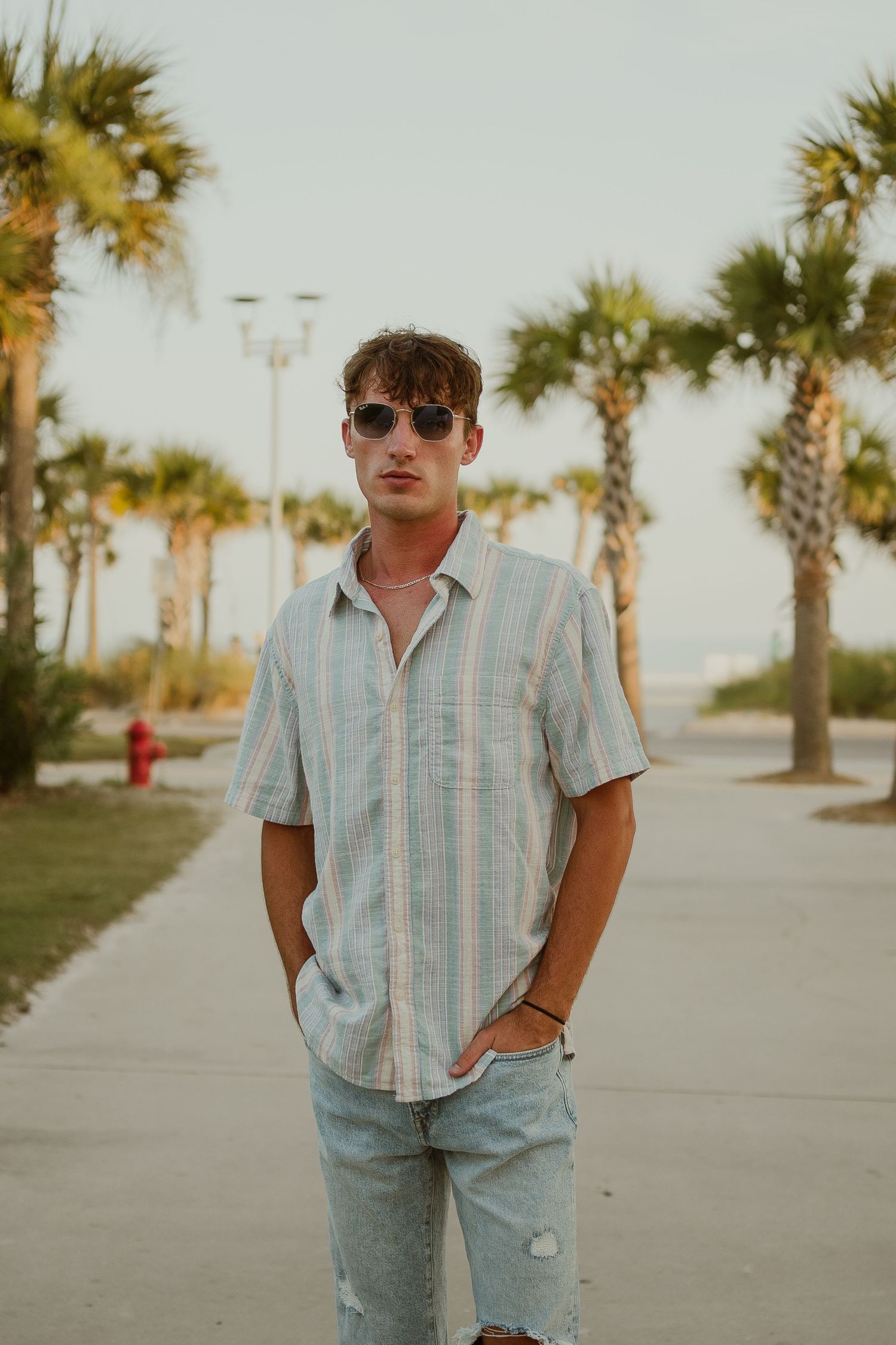 Person in striped shirt and jeans poses near palm trees at sunset.
