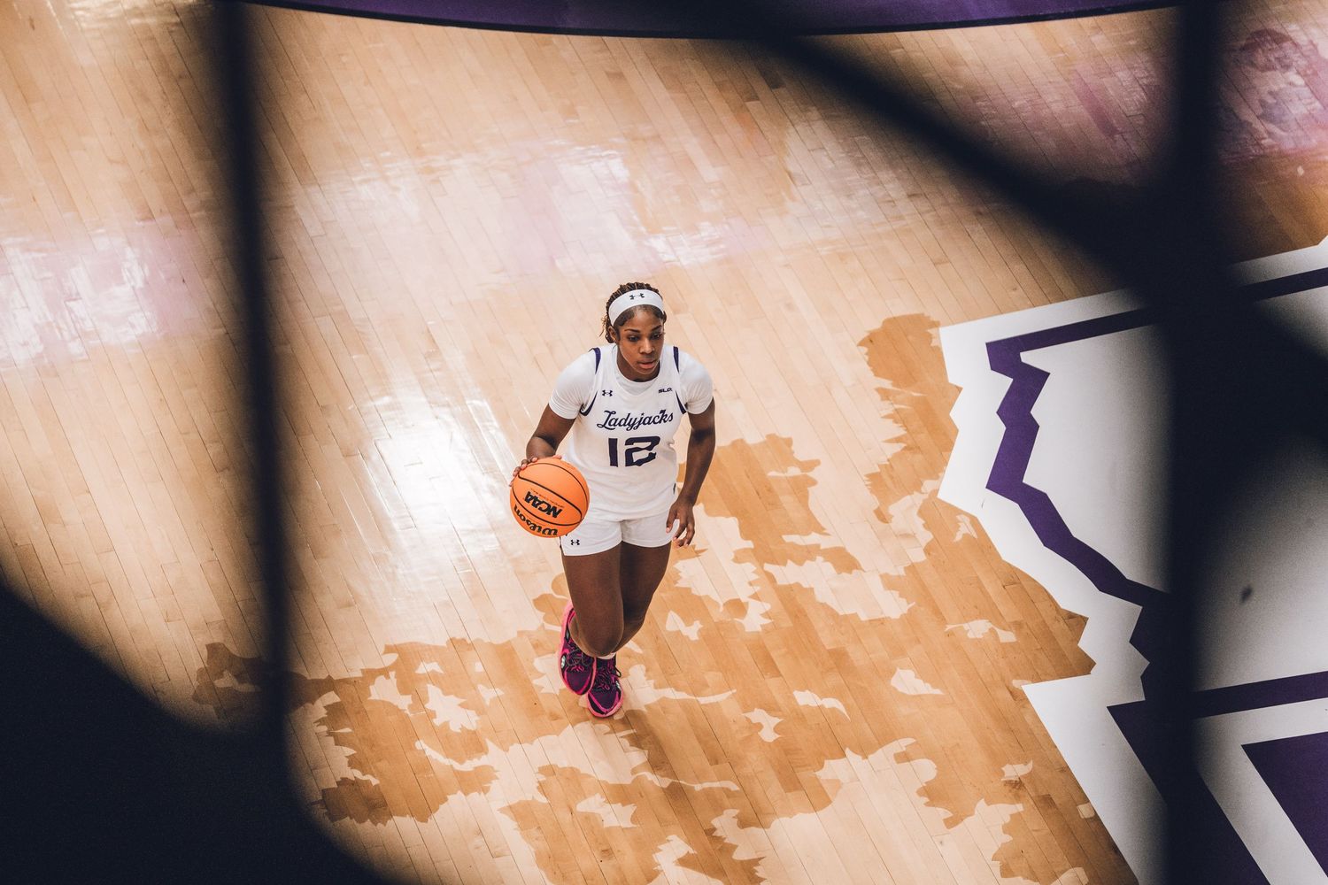 Female basketball player wearing number 12 jersey dribbles on a hardwood court, viewed from above through a basketball hoop.