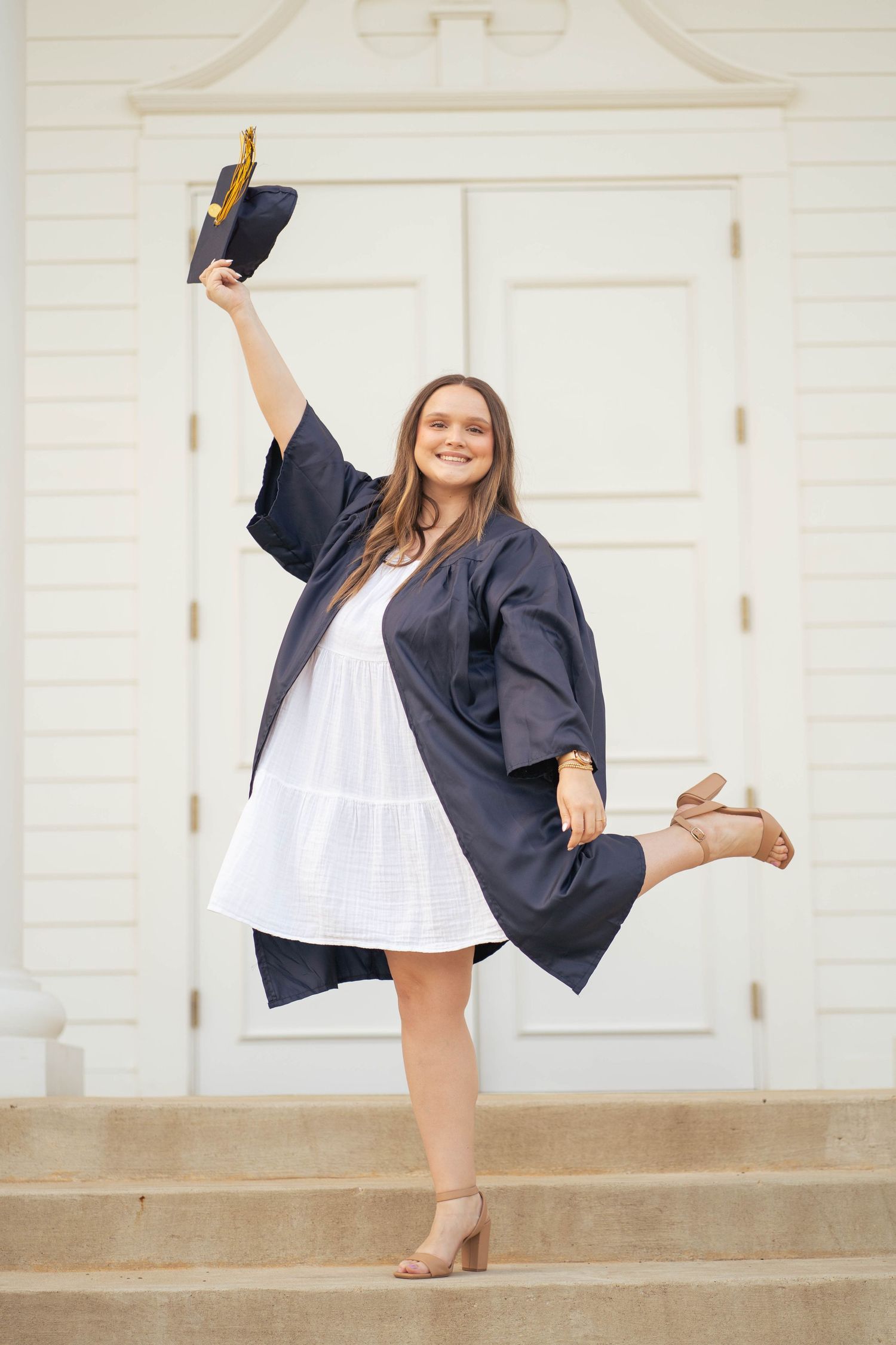 Graduate in navy cap and gown celebrates on steps wearing white dress and tan heels.