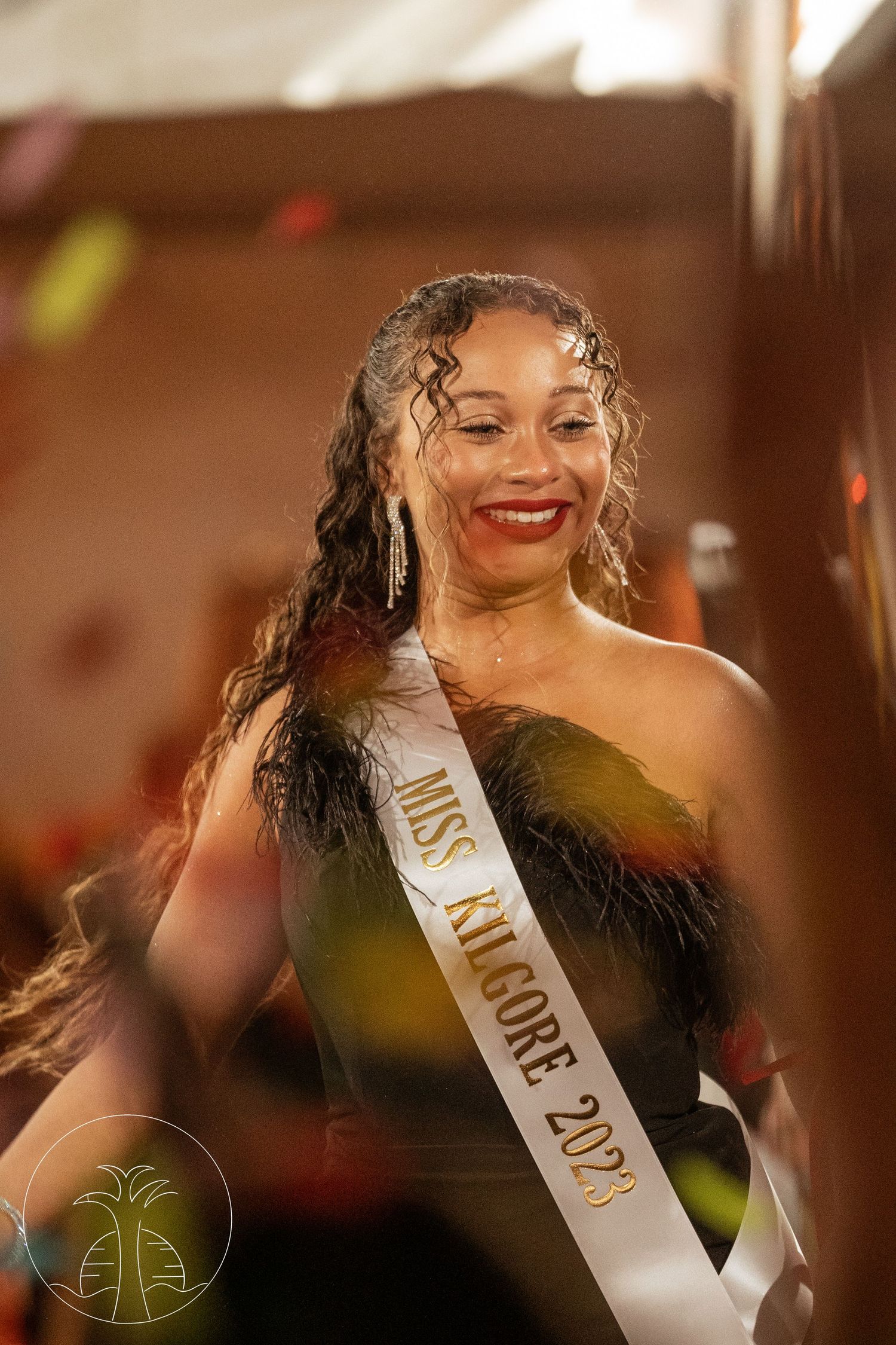 Beauty pageant contestant wearing sash and elegant black formal dress smiles on stage.