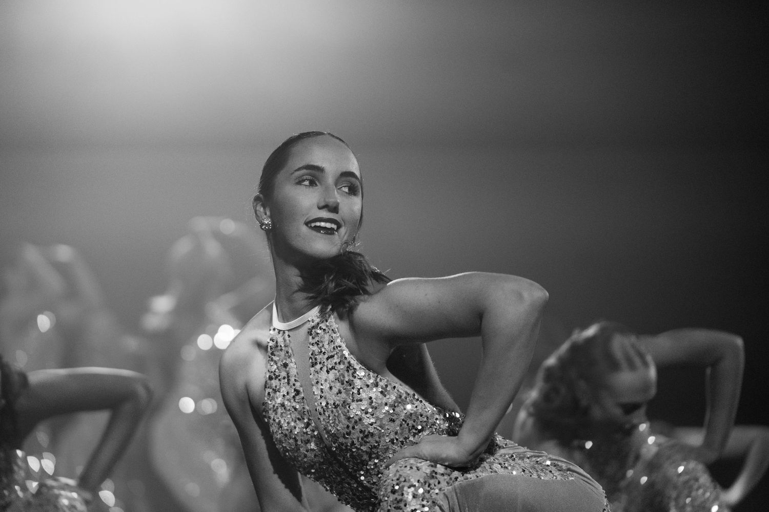 A young female dancer in a sequined costume performs on stage under dramatic lighting in black and white.