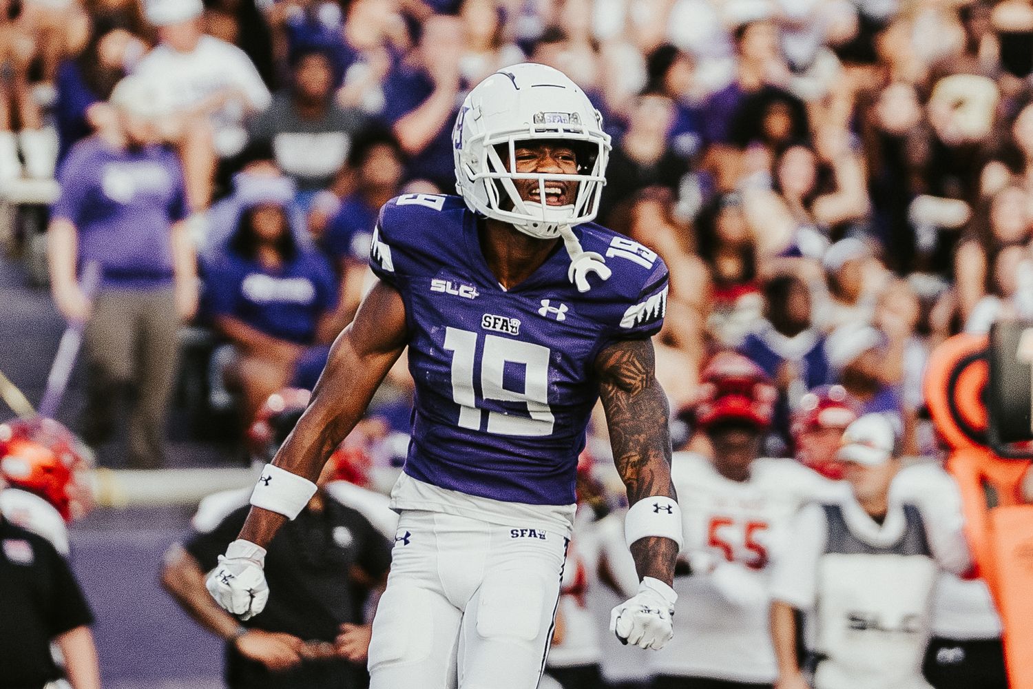 A college football player in purple and white uniform carries the ball during a game against opposing team.