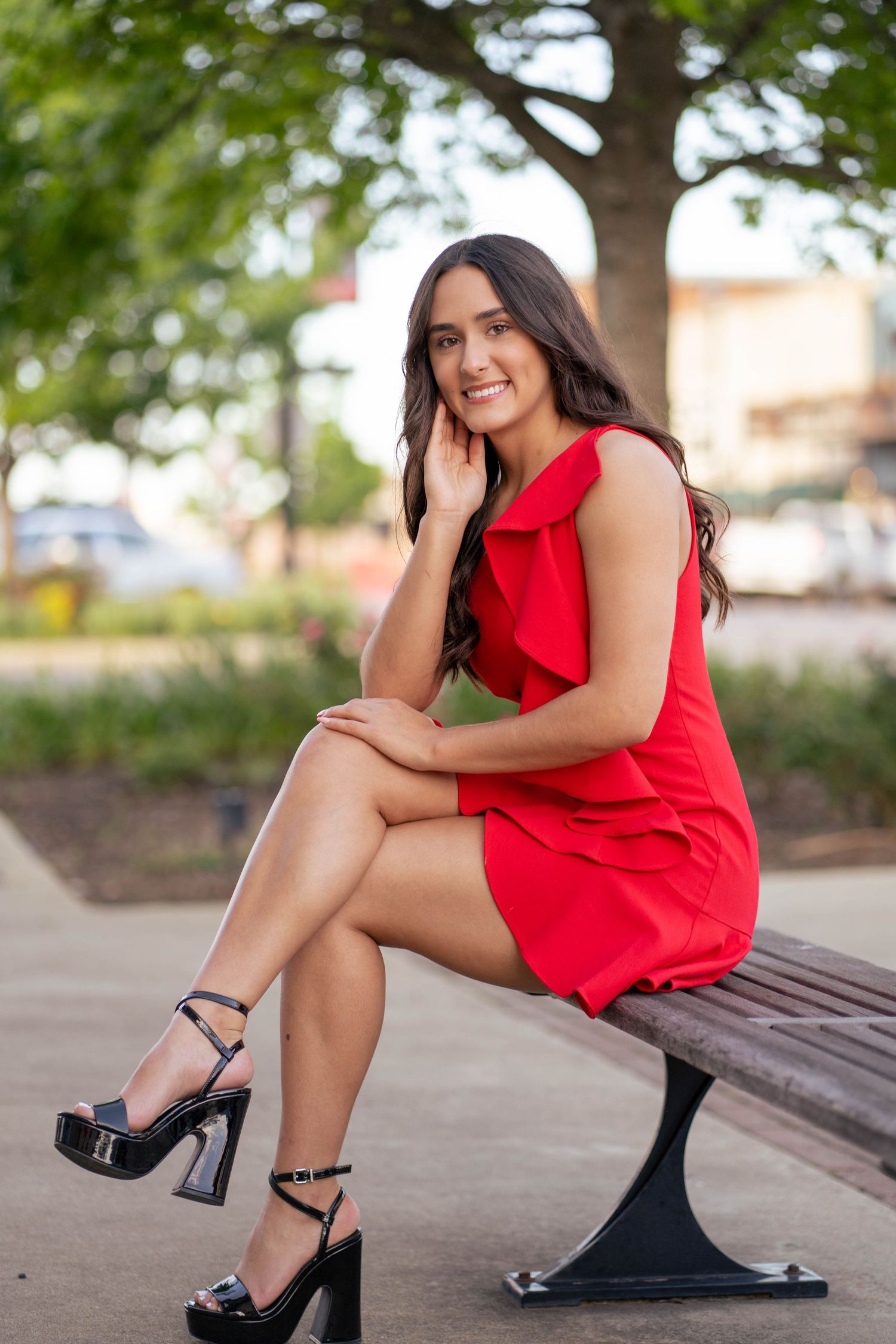 Woman in red dress and heels poses elegantly on outdoor bench during photo session.