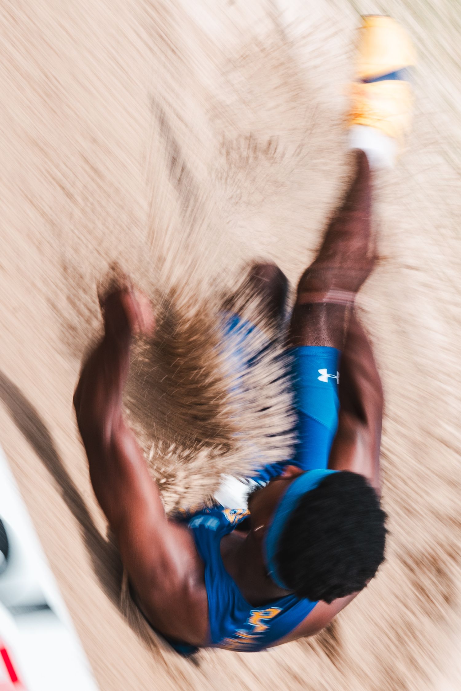 Aerial view of athlete in blue Under Armour uniform landing in sand pit during long jump competition.