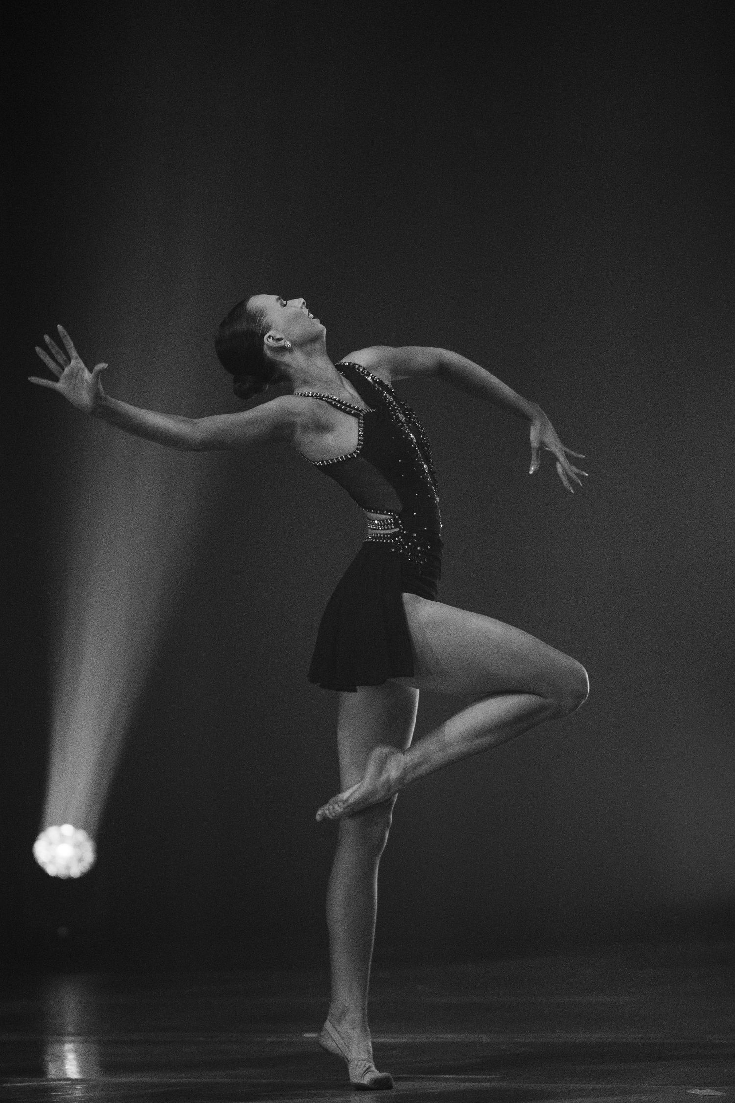 Black and white photo of a female dancer performing on stage, balancing on one foot under a spotlight.