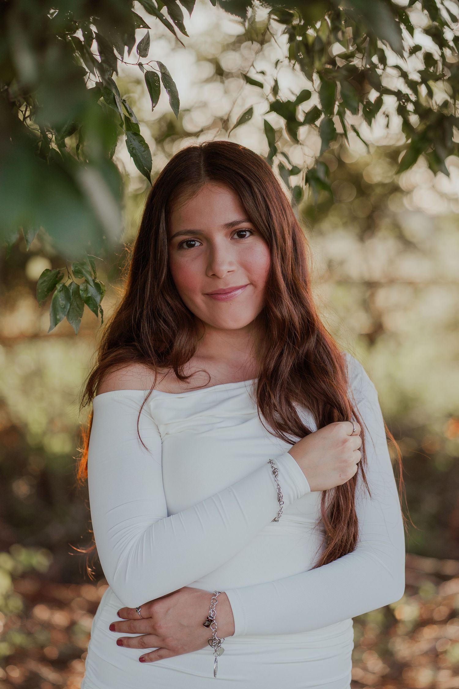Portrait of woman in white off-shoulder dress posing under tree branches in natural lighting.