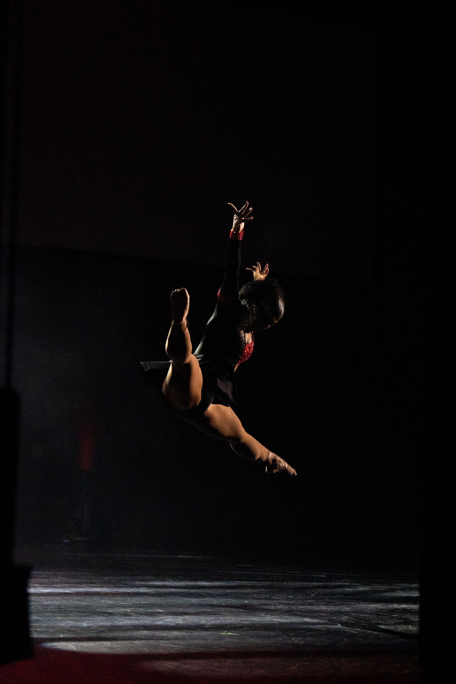 Gymnast performing a dramatic mid-air leap in a dark studio, illuminated by a single spotlight.