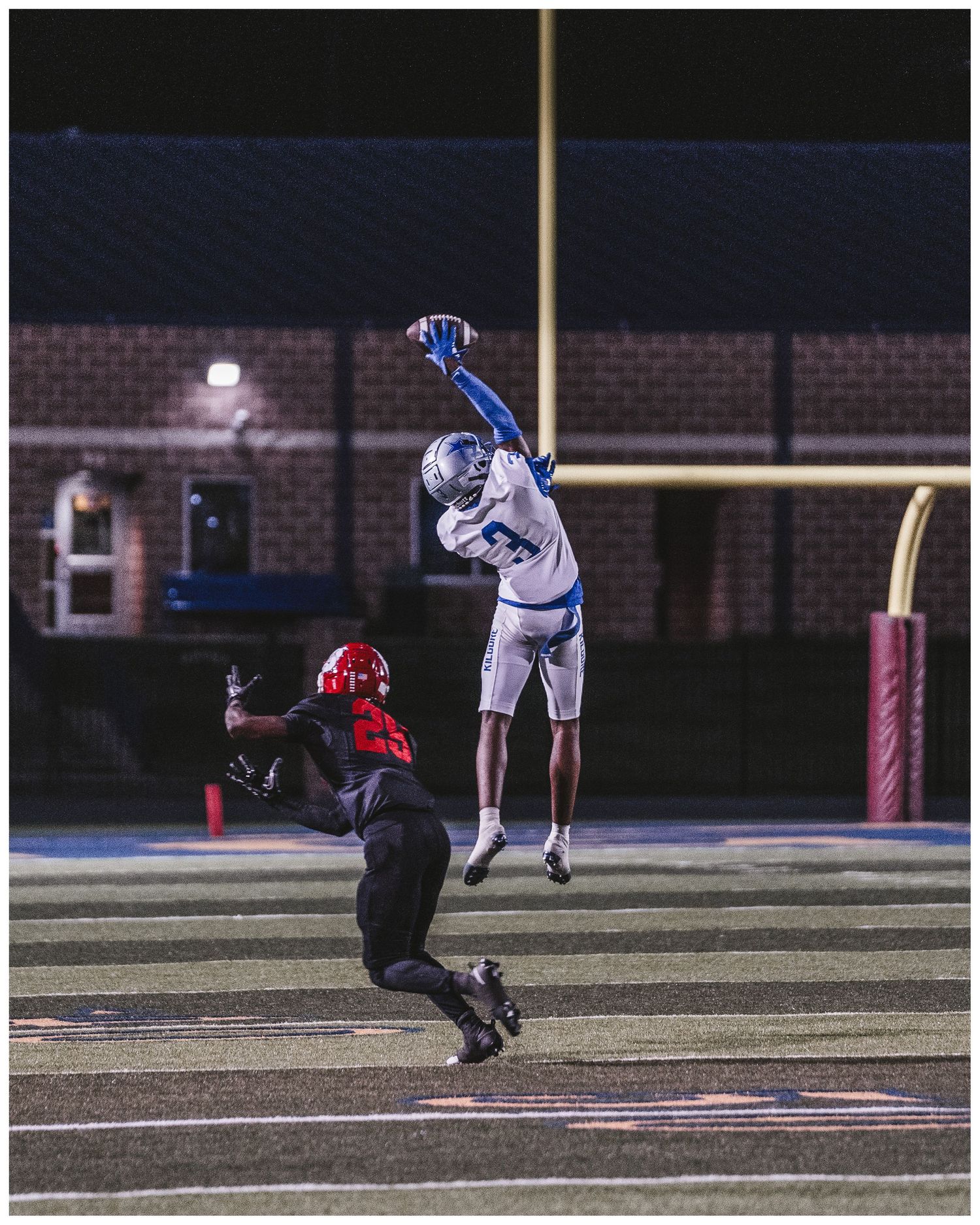 Two football players compete for a pass during a nighttime game under stadium lights on a brick-walled field.