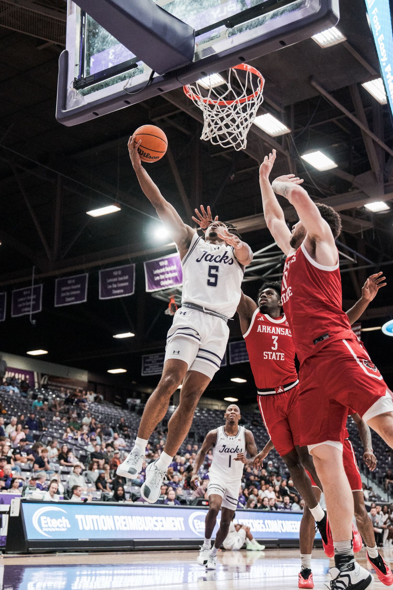 Basketball player in white jersey drives to the hoop against defenders in red uniforms during intense game action.