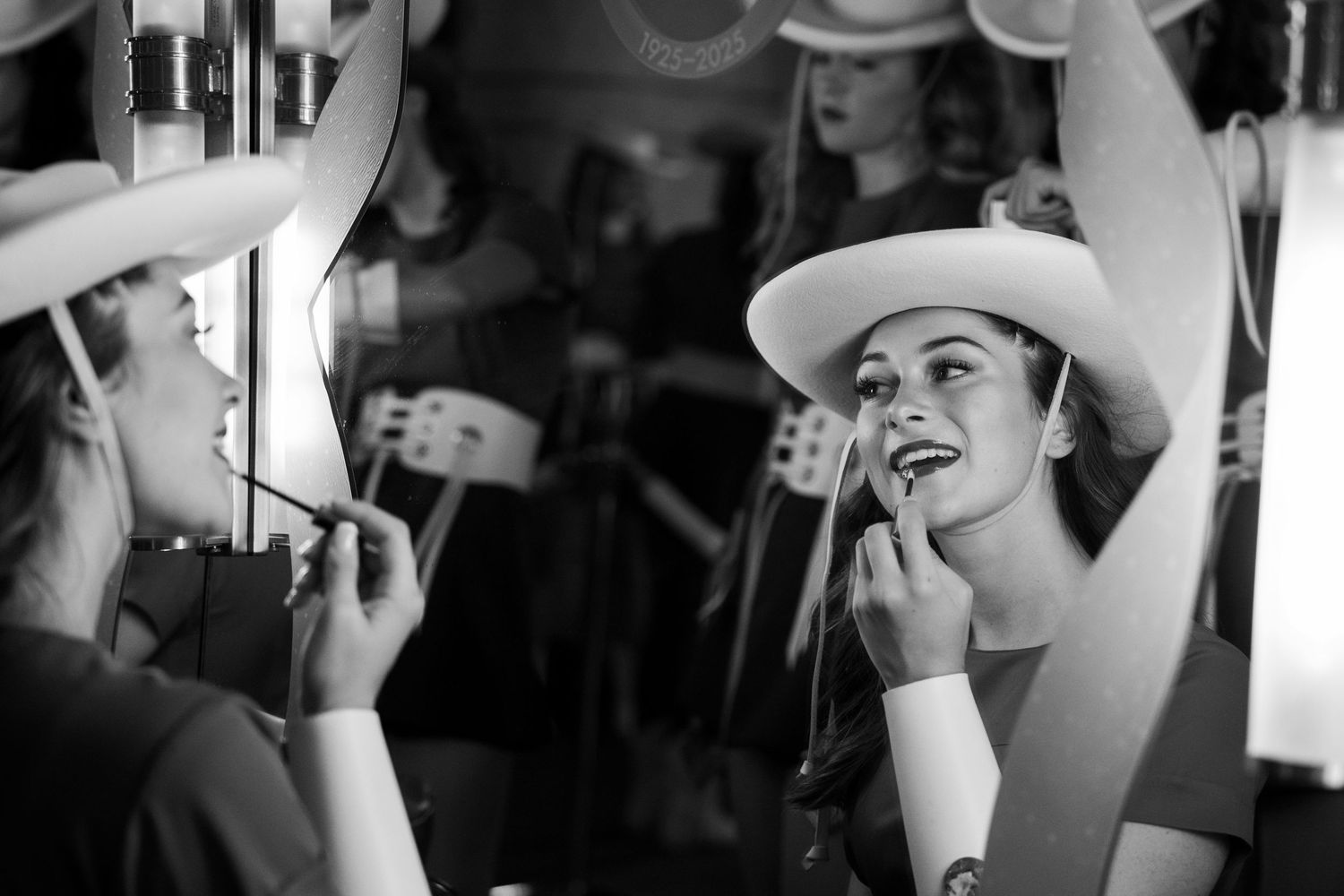 A person in a cowboy hat laughs joyfully while applying makeup in a mirror reflection.