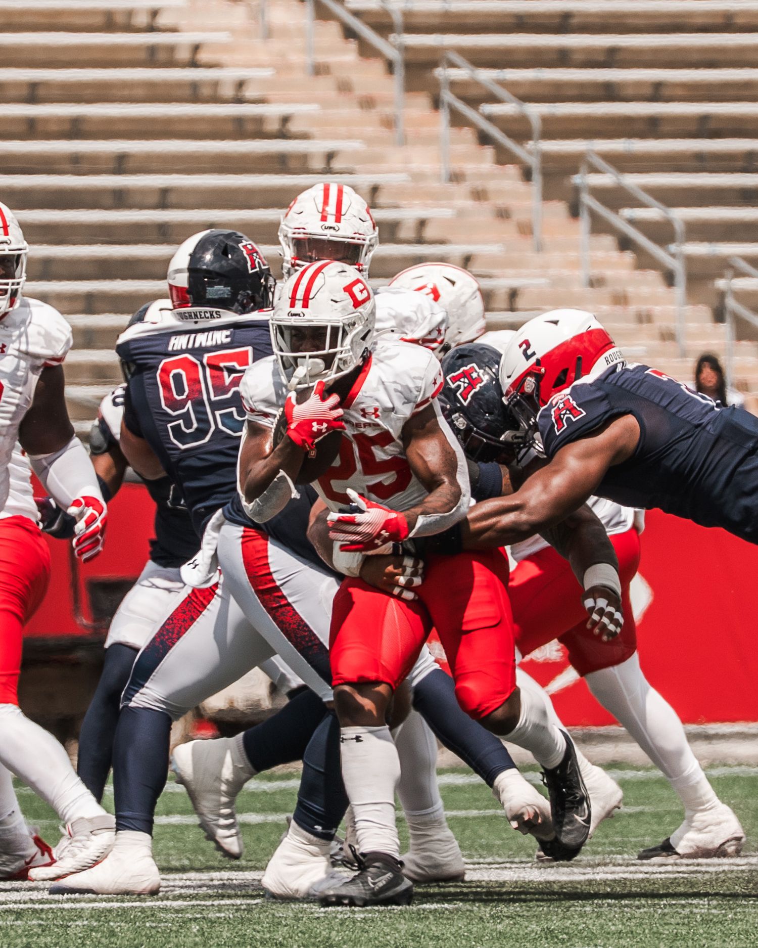 College football players in red and white uniforms compete in a tackle during a daytime game in a stadium.
