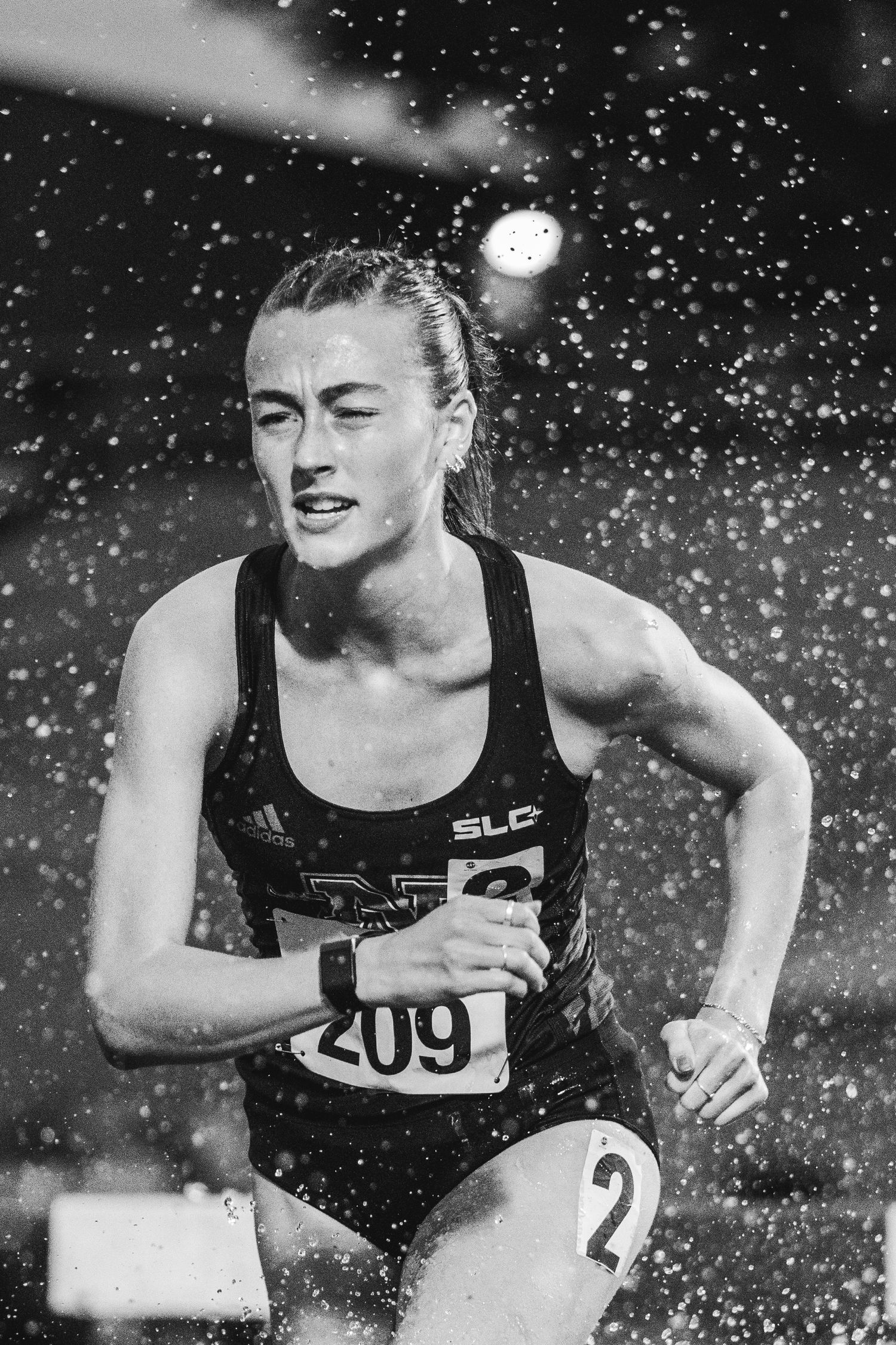 Female athlete number 209 races intensely through water spray in a black and white steeplechase competition photo.