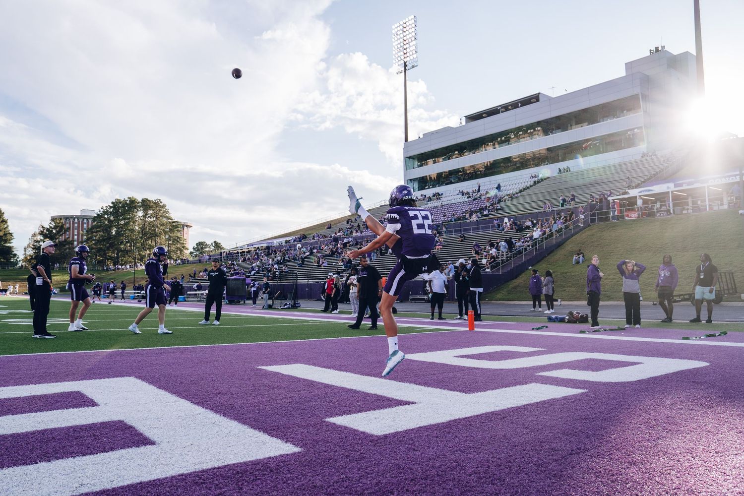 Football players leap for the ball in the endzone during a game at a stadium with purple and white field markings.