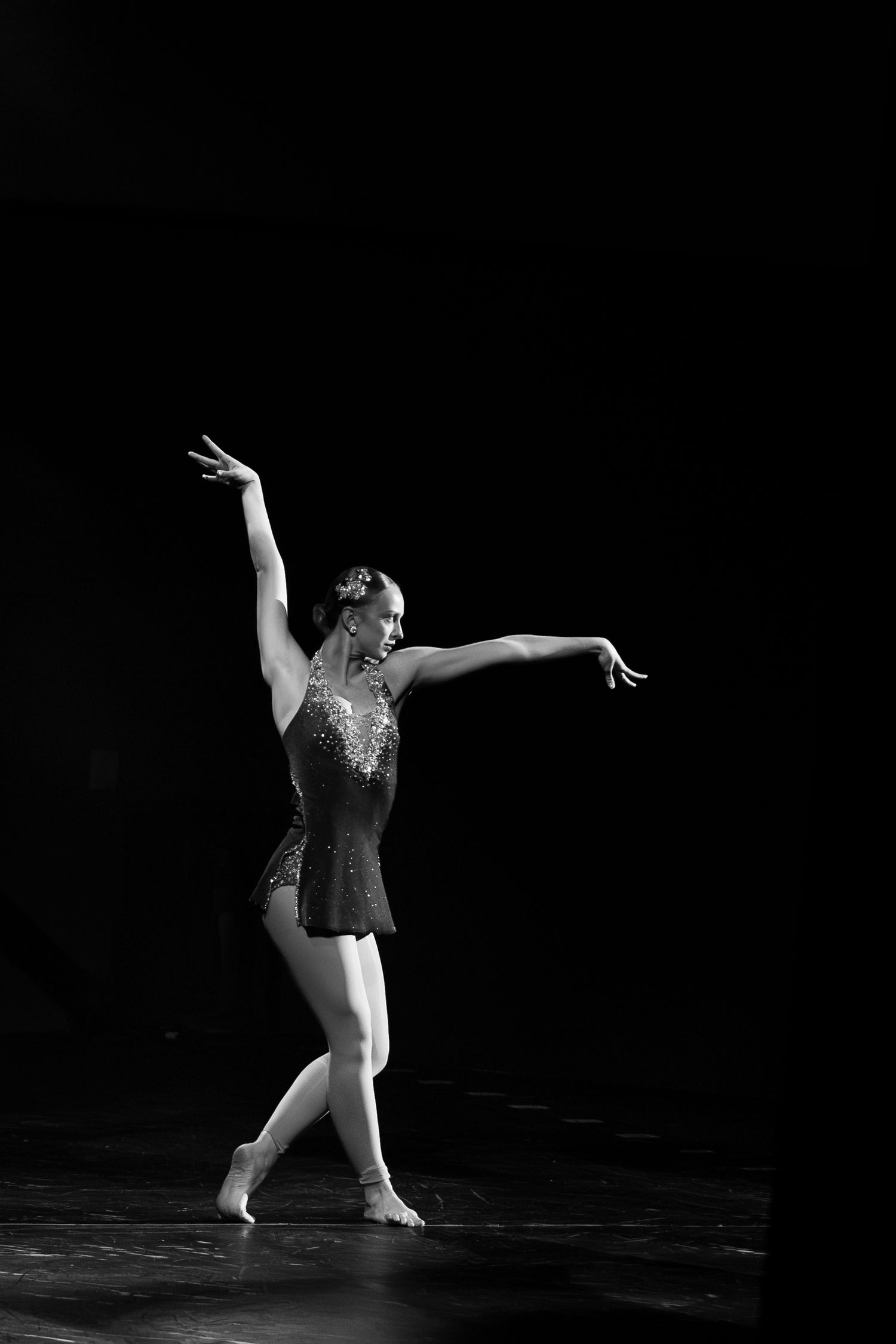 Black and white photo of a ballet dancer performing on stage in a sequined dress with arms gracefully extended.