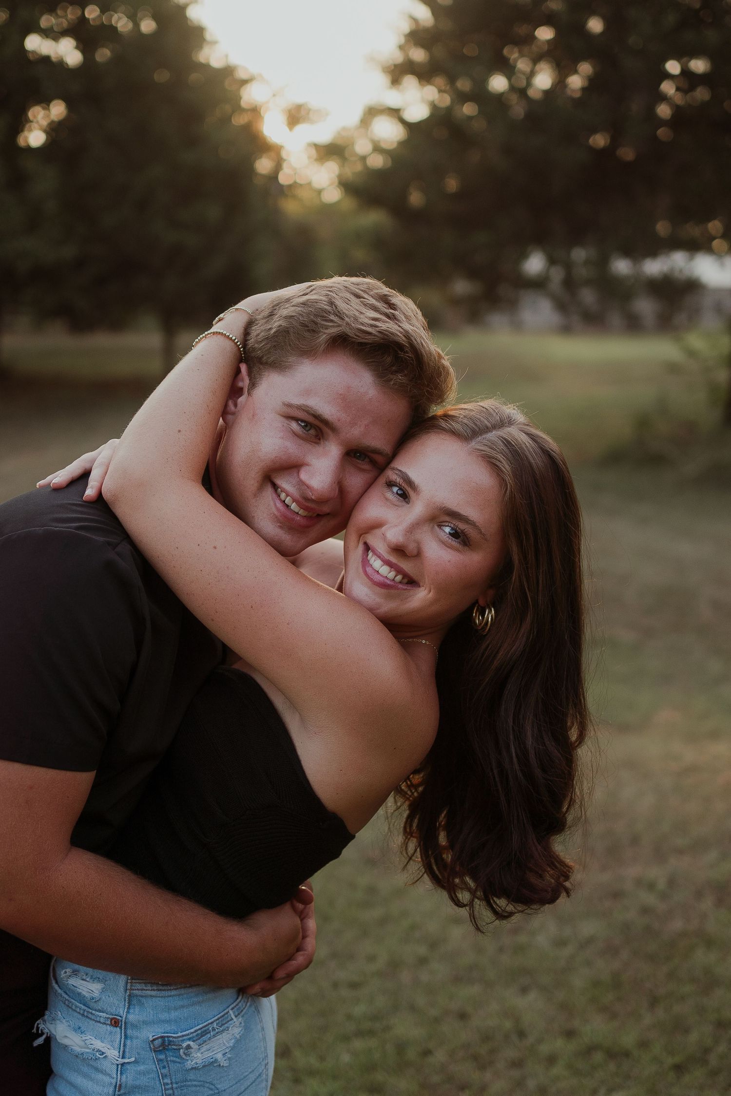 Playful couple in casual wear having fun while embracing on grassy field at sunset.