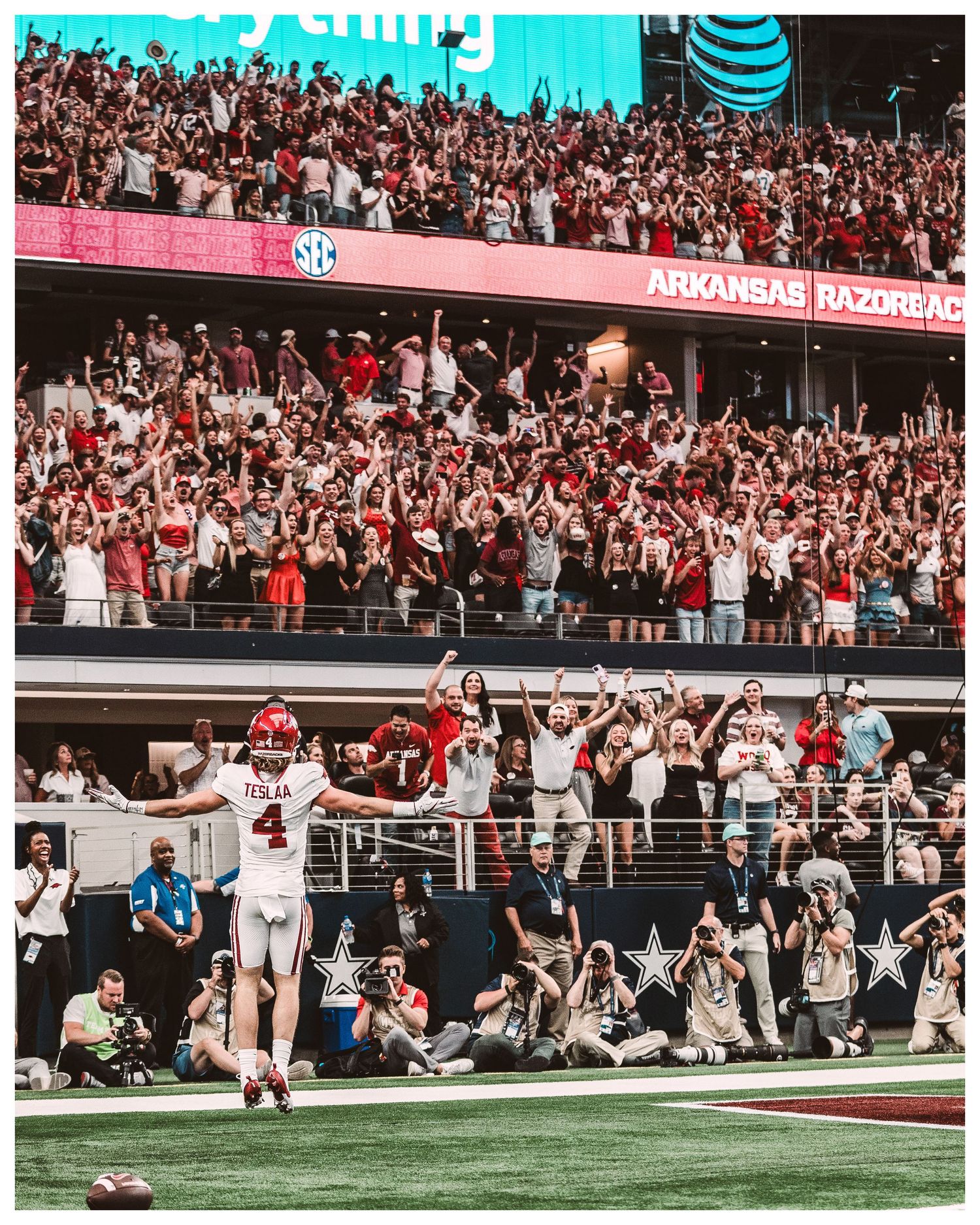 Football player celebrates with enthusiastic fans in packed stadium stands during a game with vibrant blue sky overhead.