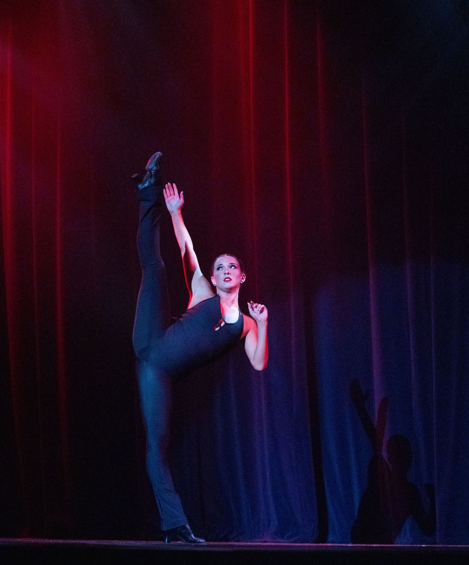 A flexible female dancer performs a high leg extension on a dramatic red and blue lit stage.