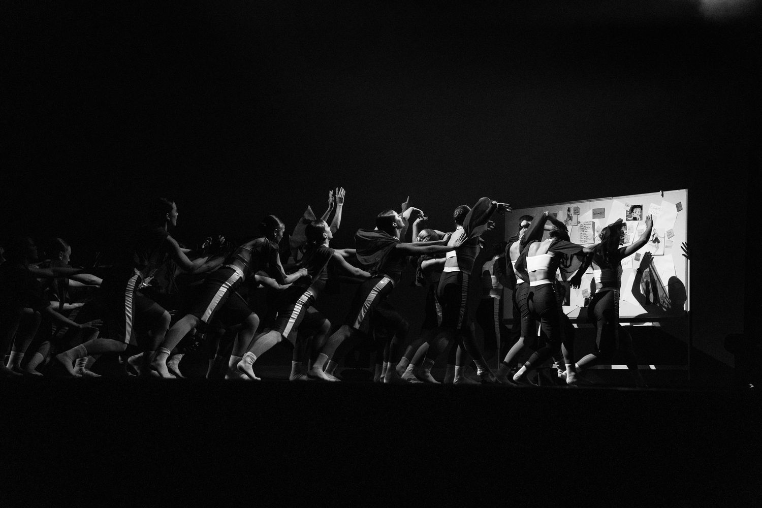 Black and white photo of dancers performing on a dark stage with a illuminated screen backdrop.