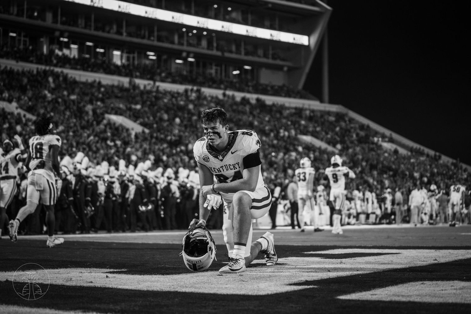 Black and white photo of a football player kneeling on the field at night, helmet on ground, stadium crowd in background.