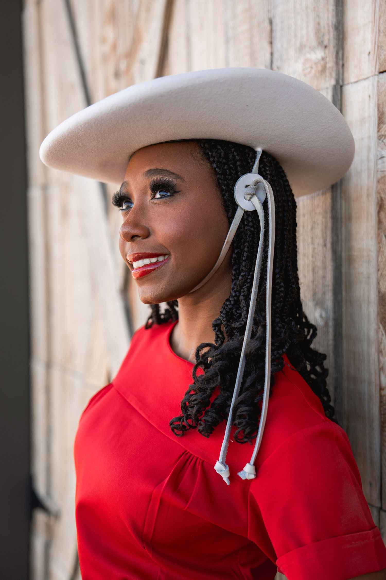 A person in a red coat and white cowboy hat smiles while looking to the side against a light-colored background.