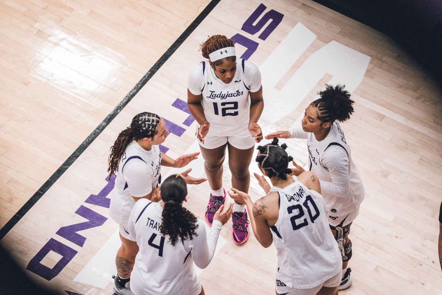 Women's basketball team huddles on court, players in white uniforms with numbers 4, 12, and 20 visible.