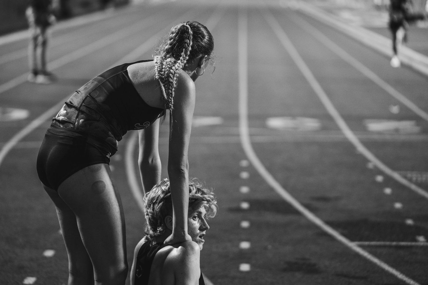 Two female athletes on a running track, one bending over catching her breath after a race in black and white.