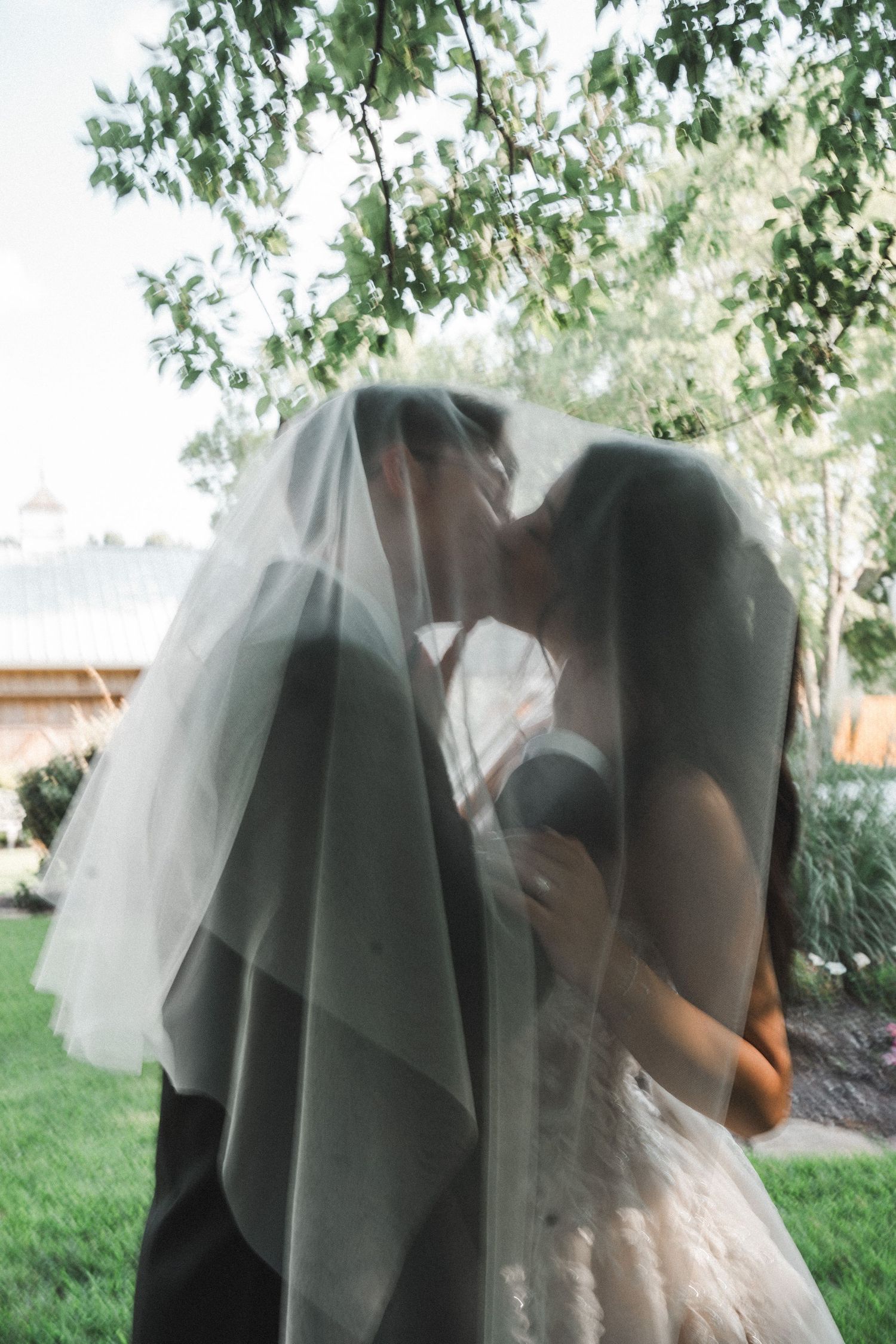 Newlyweds share an intimate kiss under a gossamer veil in an outdoor garden setting.