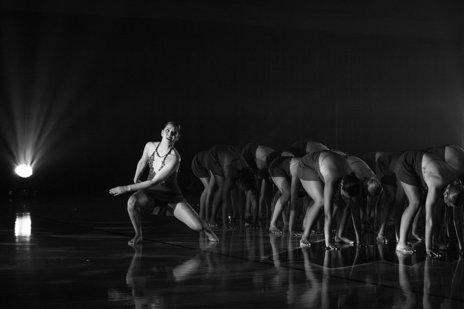Black and white photo of dancers performing in dramatic formation with spotlight illumination.