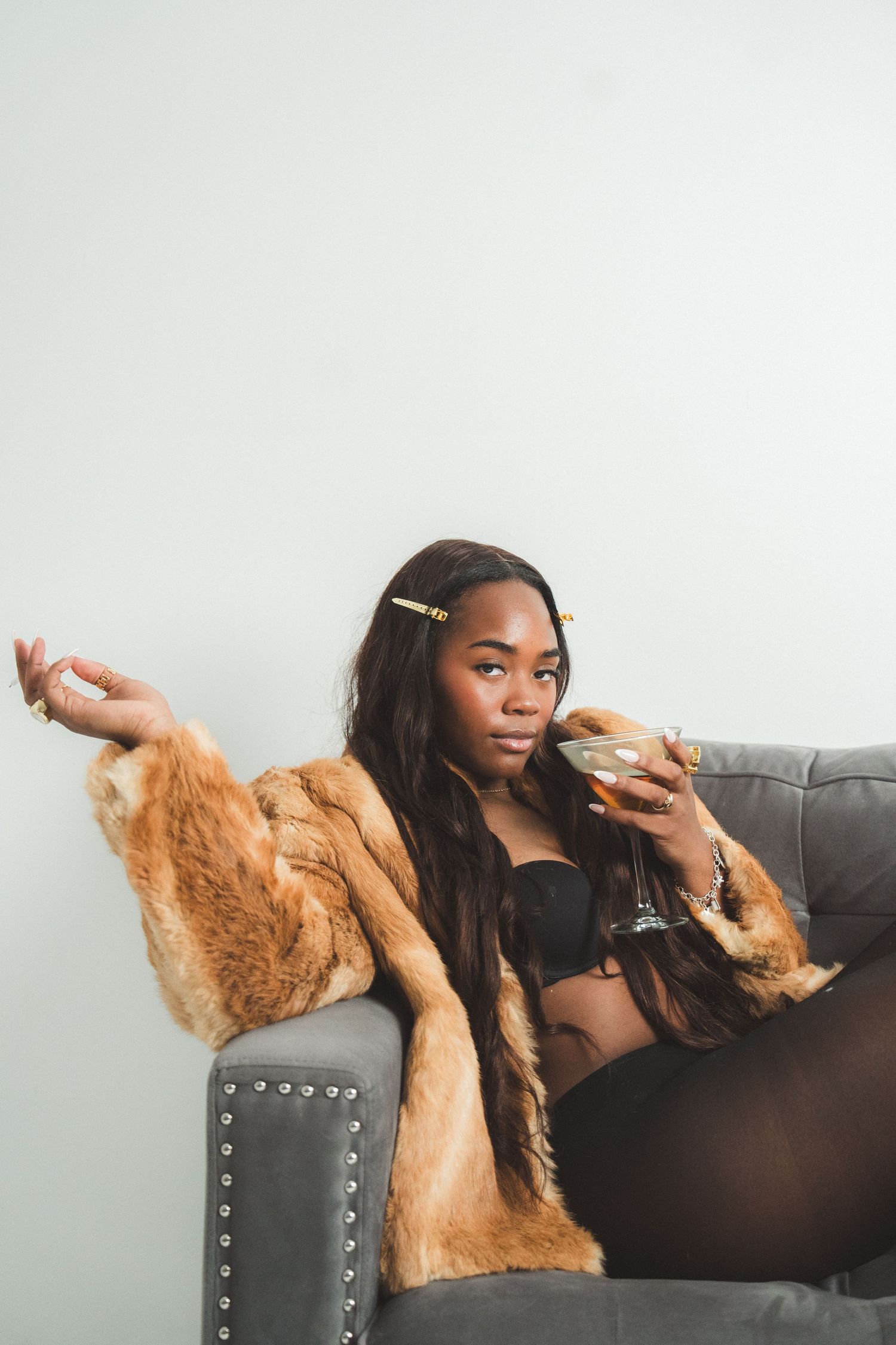 A relaxed figure lounges on a gray couch wearing a brown fur coat and black crop top in a minimalist setting.