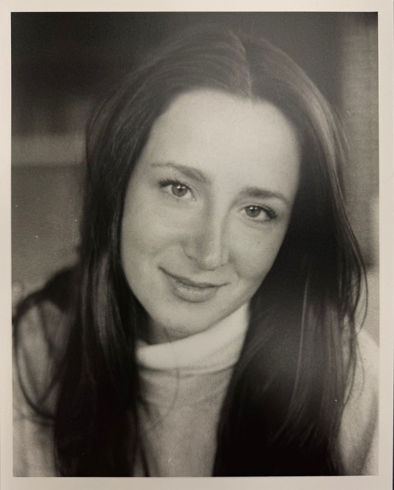 A black and white portrait photograph showing a person with long dark hair wearing a turtleneck sweater and gentle smile.