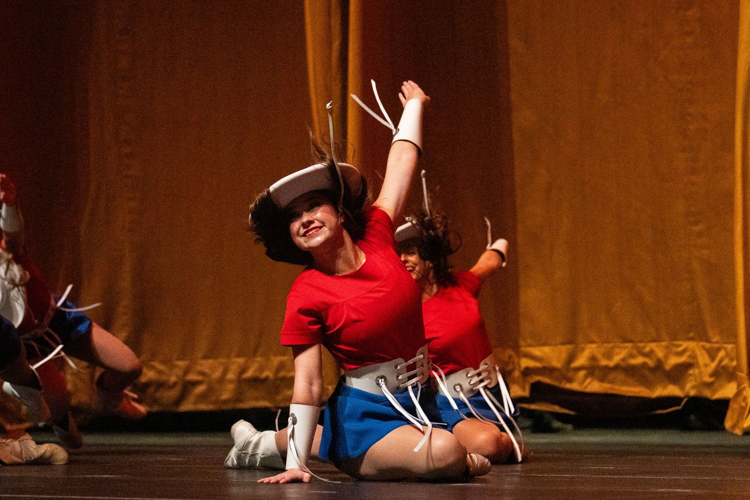 A dancer in a red top and blue shorts performs an energetic western dance routine on stage.