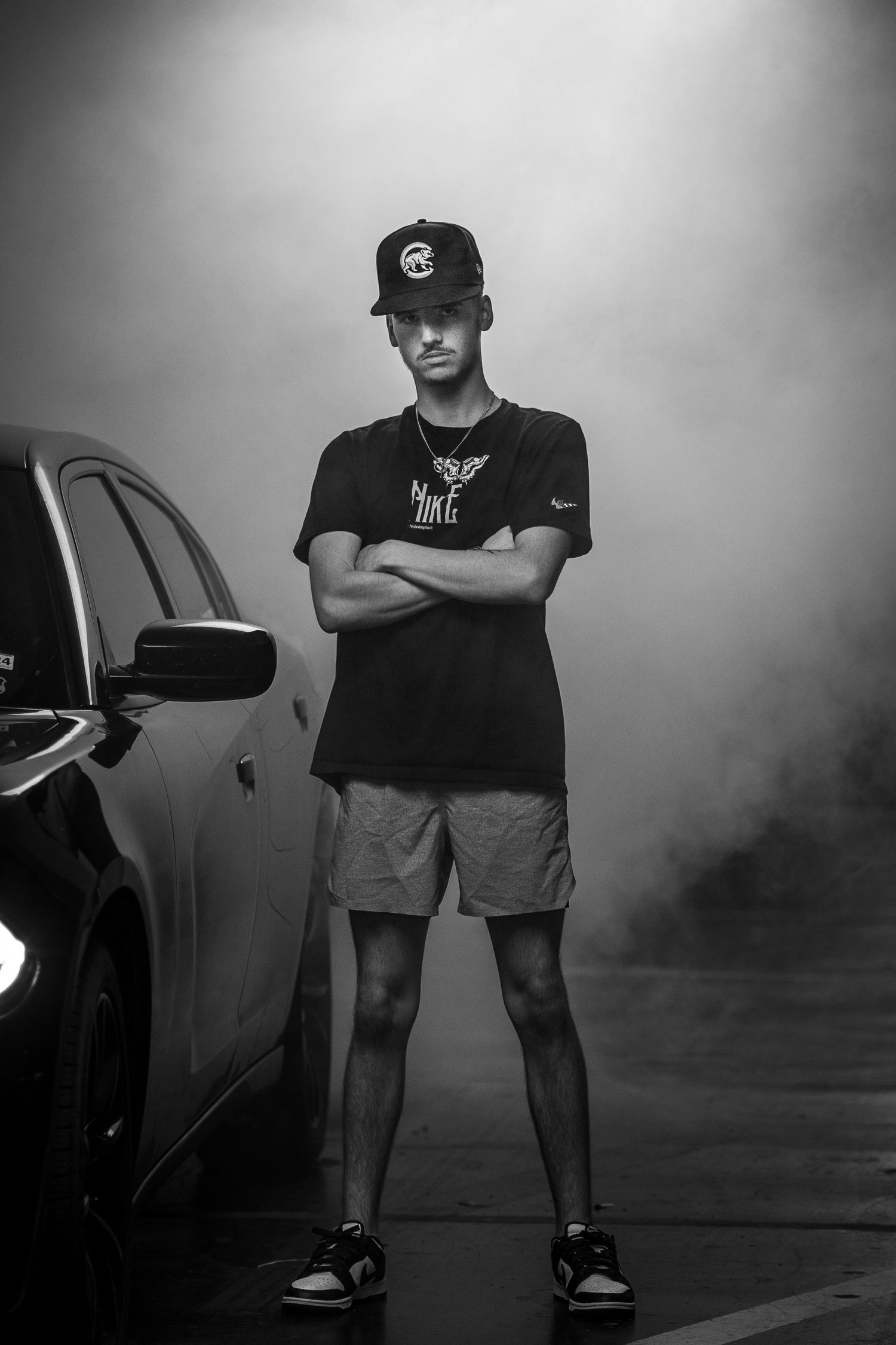 Dramatic black and white photo of person in casual wear standing beside car in fog.