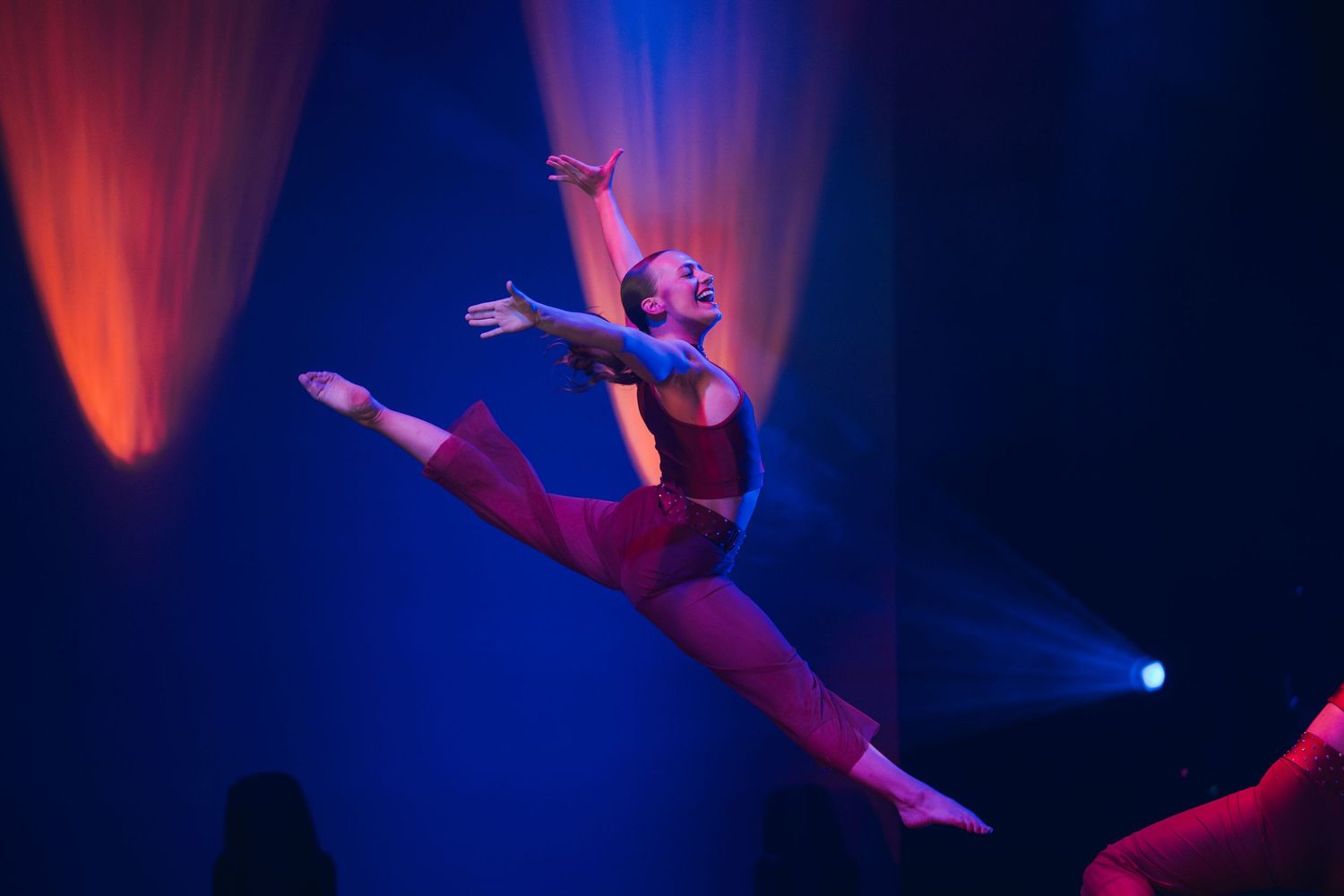 A dancer performs dramatic splits in red stage lighting against a dark blue background during a theatrical performance.