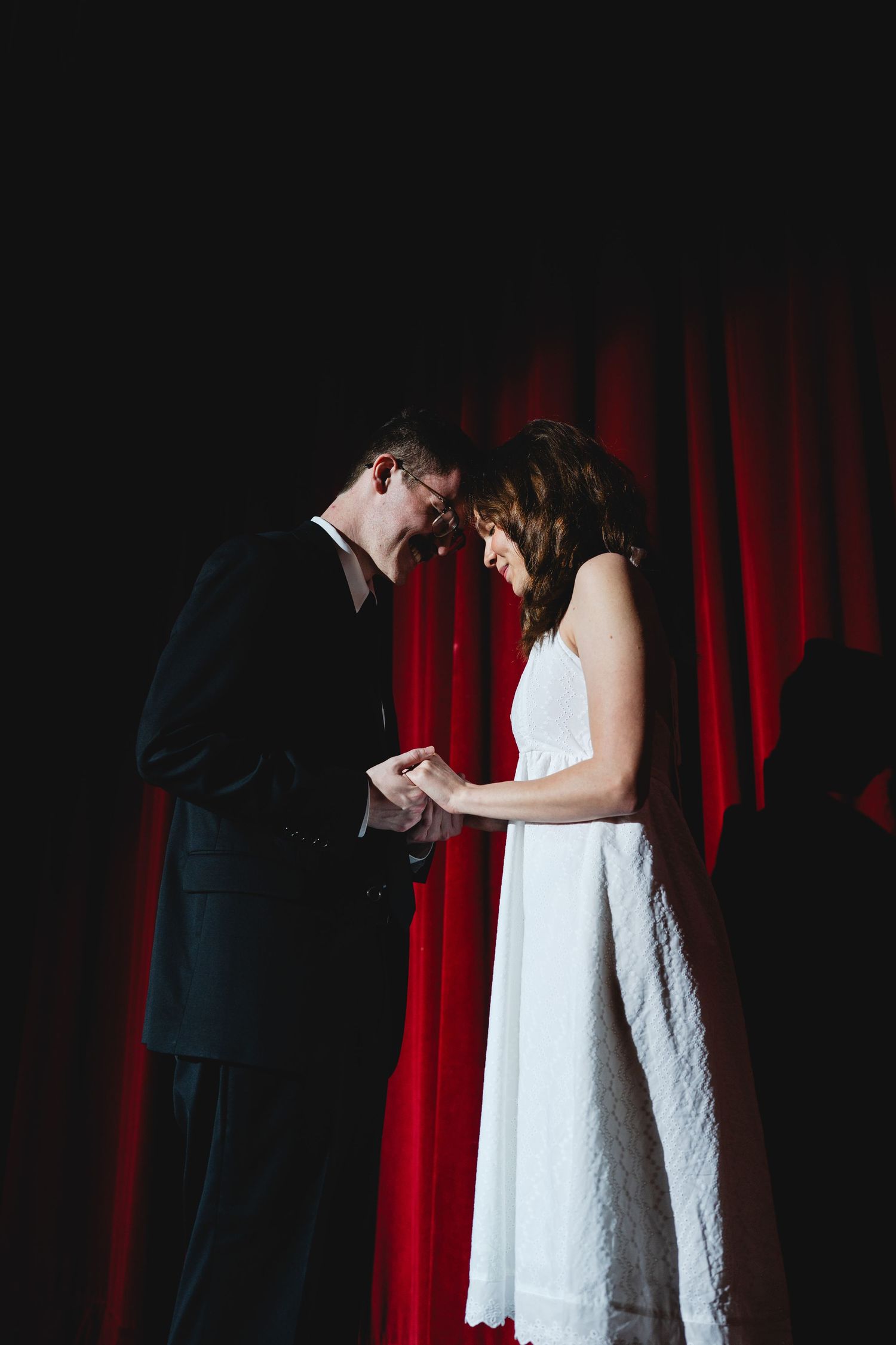 Bride and groom exchange rings on stage before red curtain in intimate wedding ceremony.