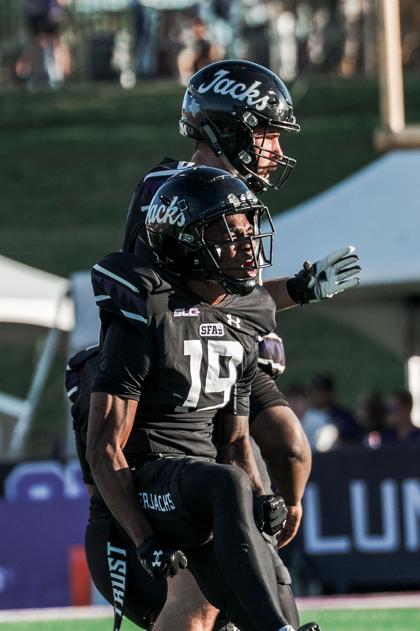 Football players in black uniforms and helmets signal plays during a game on a sunny field.