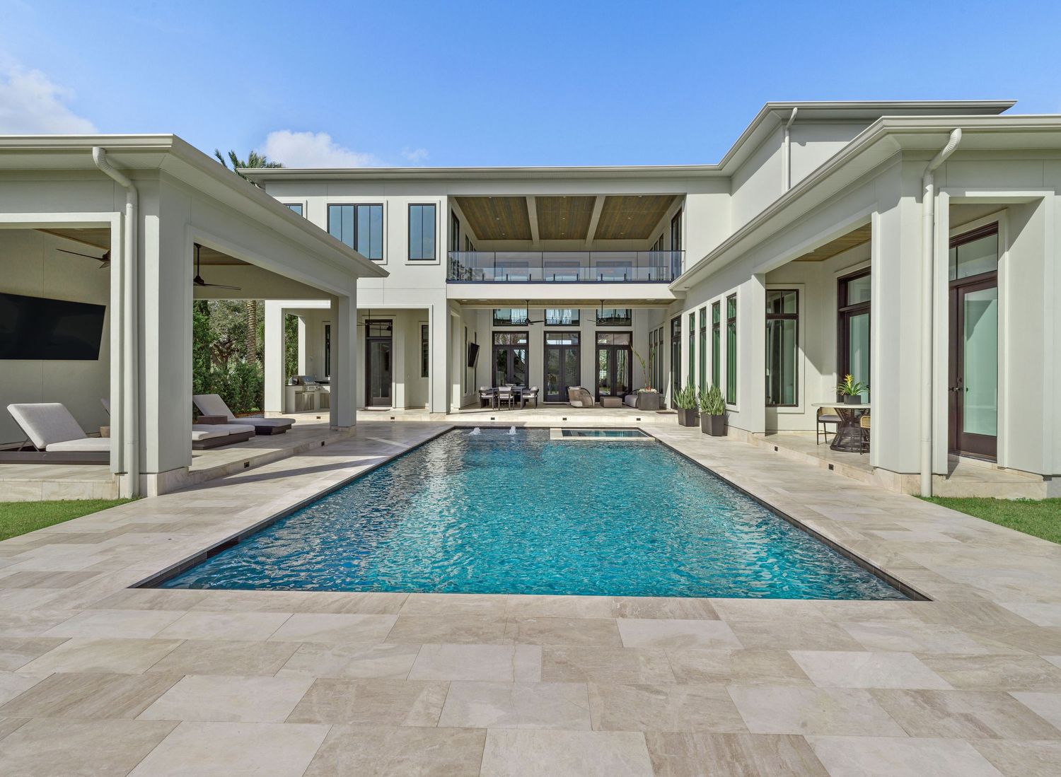 Long rectangular swimming pool adjacent to white modern home with covered outdoor living space.