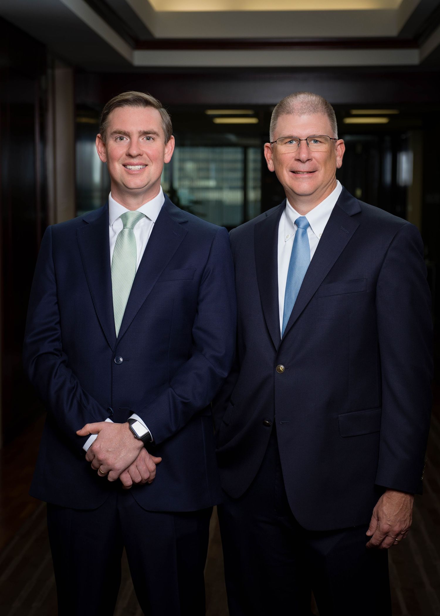 Two executives in business suits pose together for corporate portrait in office hallway.