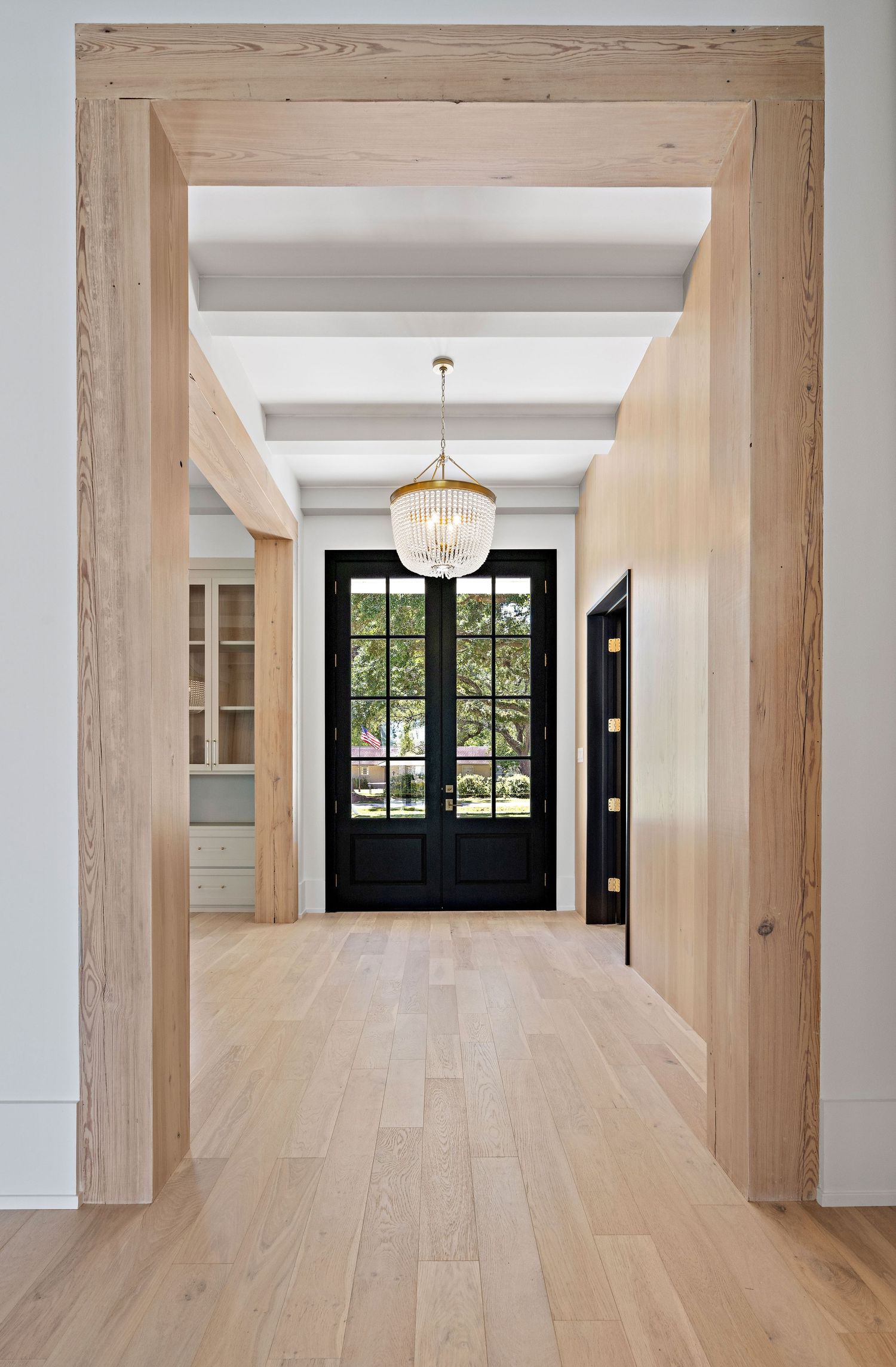 Modern entryway features light wood floors, white beams, and a black double door illuminated by an elegant crystal chandelier.