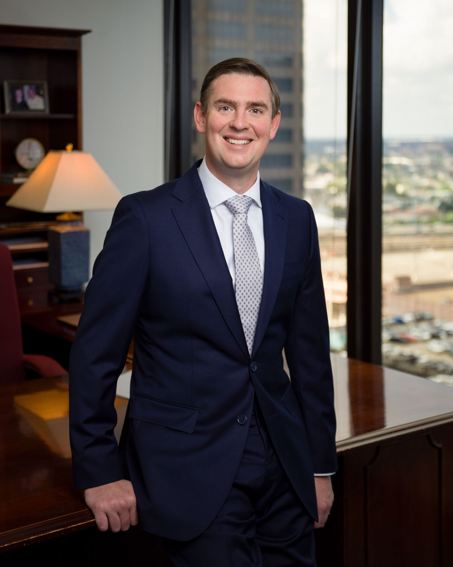 Professional business portrait of smiling executive in office with city view behind.