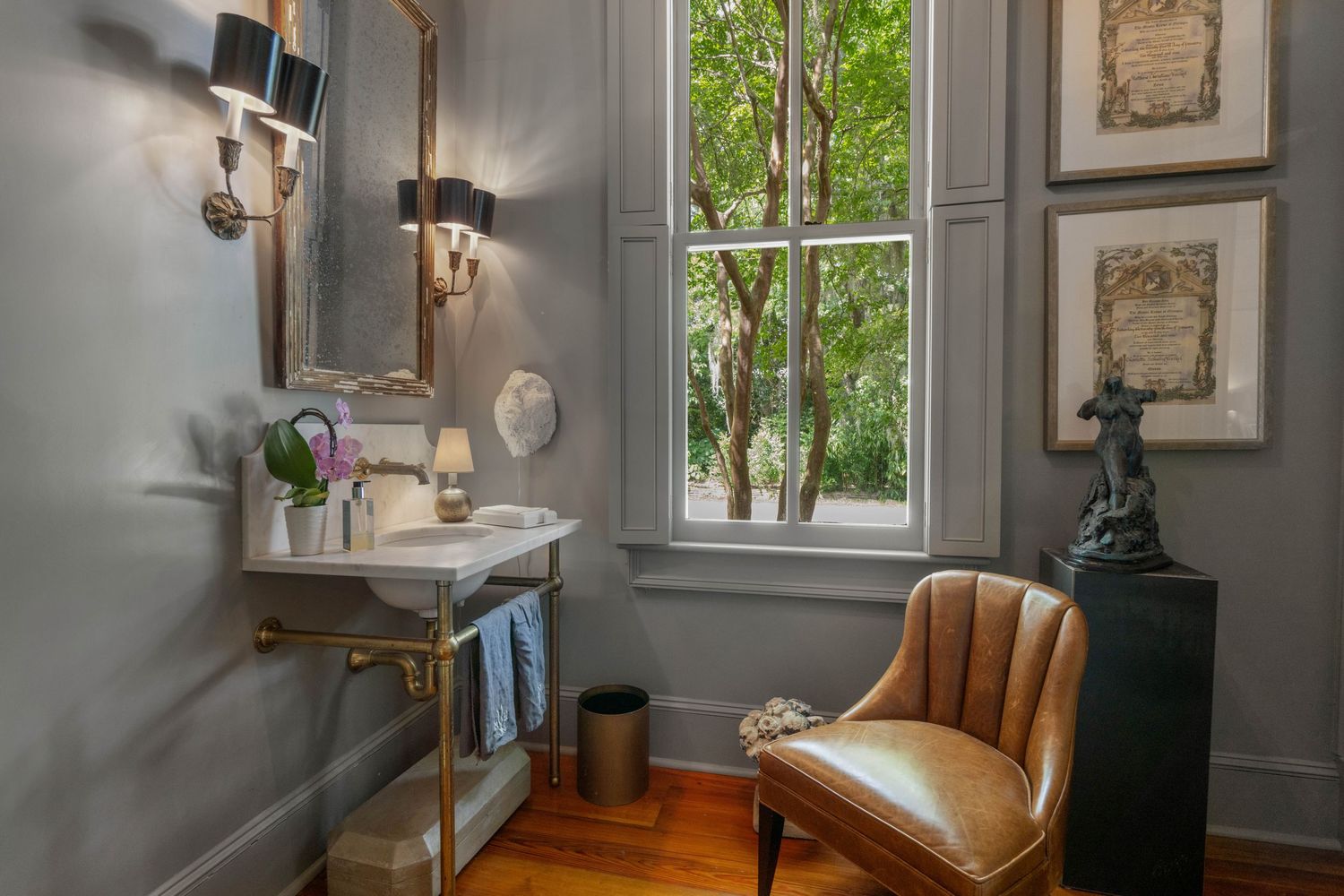 Elegant powder room with brass fixtures, vintage-style sink, and leather accent chair by tall window.