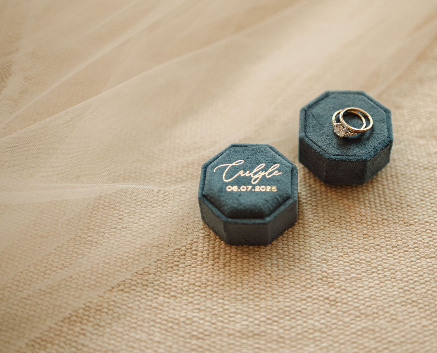 Two dark teal velvet ring boxes with gold lettering placed on beige linen fabric background.