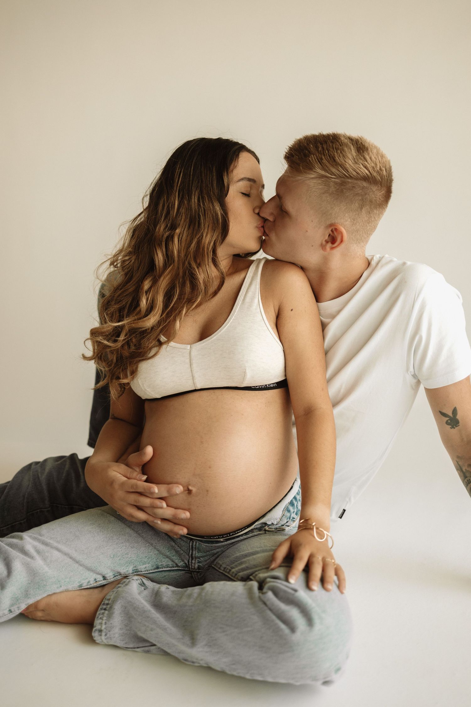 Intimate pregnancy photo series showing affectionate poses and bare baby bump with light beige backdrop.