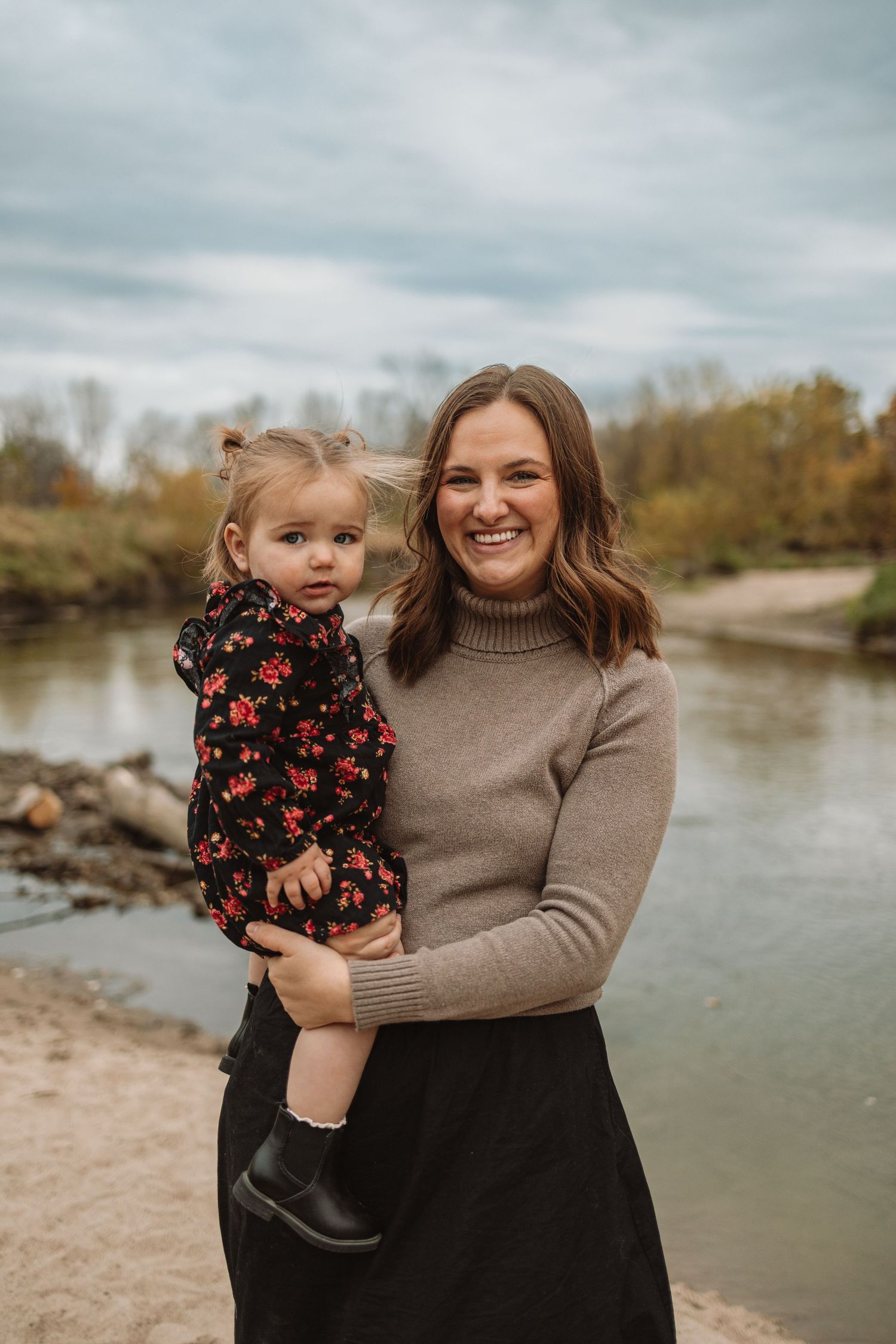 A mother and child share joyful moments together by a riverbank on an overcast autumn day.